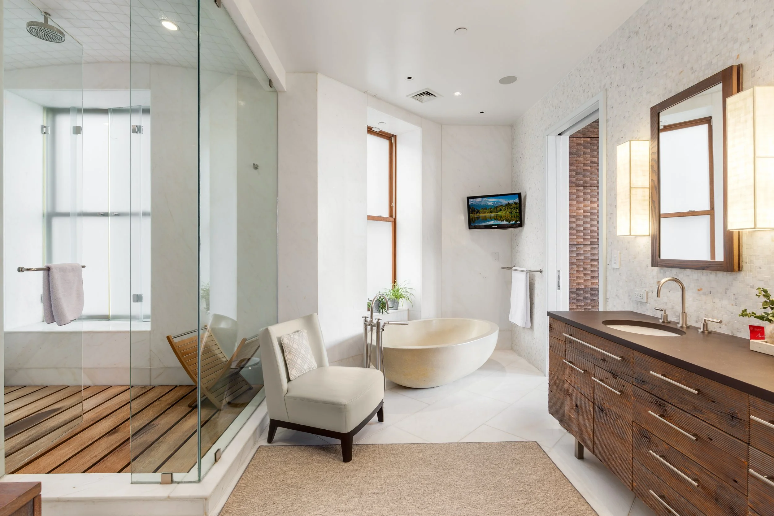 Modern bathroom with glass-enclosed shower, white armchair, freestanding bathtub, wooden vanity with sink, small wall-mounted TV, and a window with natural light.