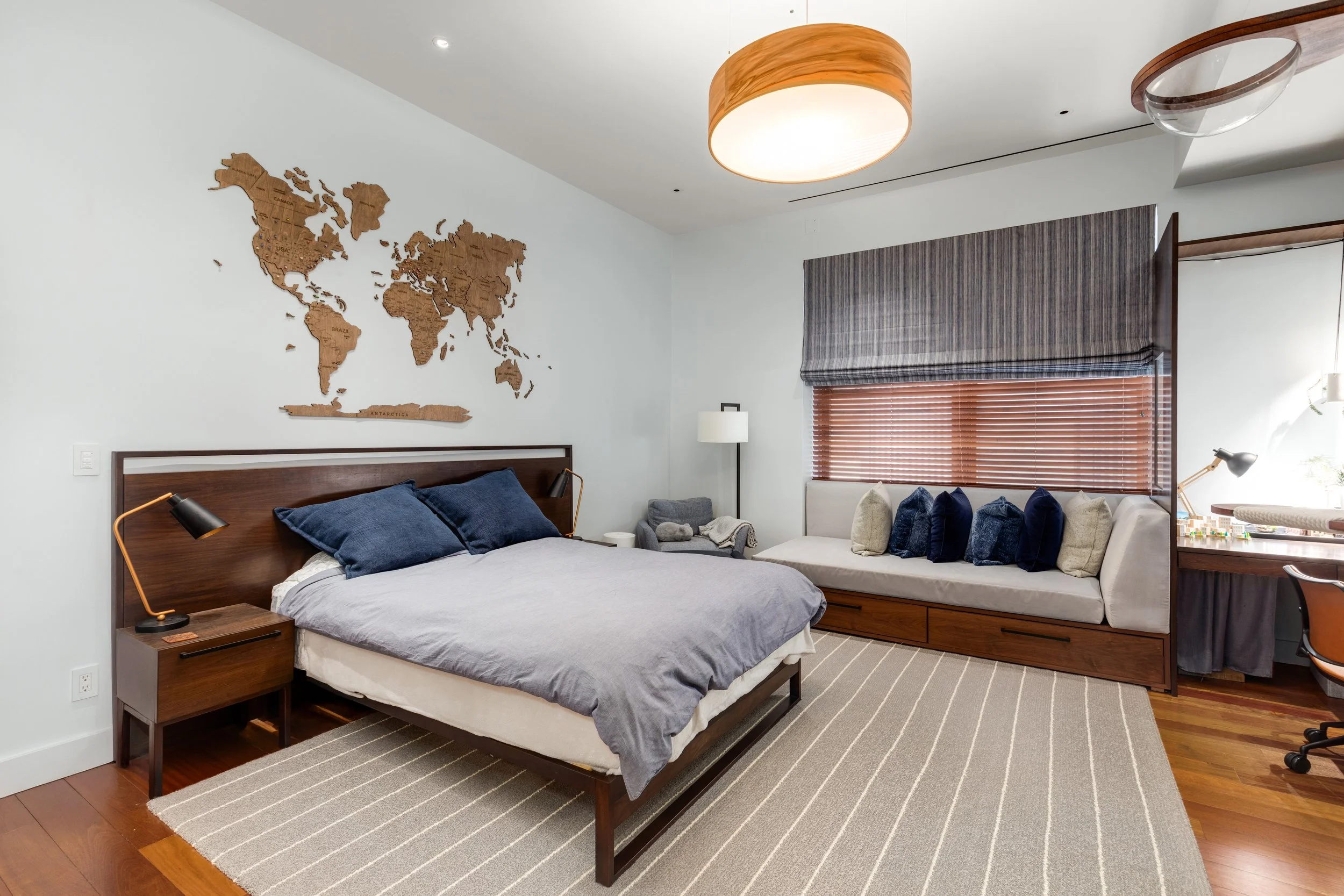 Modern bedroom with a wooden bed frame, blue pillows, and a world map wall art above it. There is a window with wooden blinds and a bench with pillows underneath. A small gray armchair and a floor lamp are also visible.