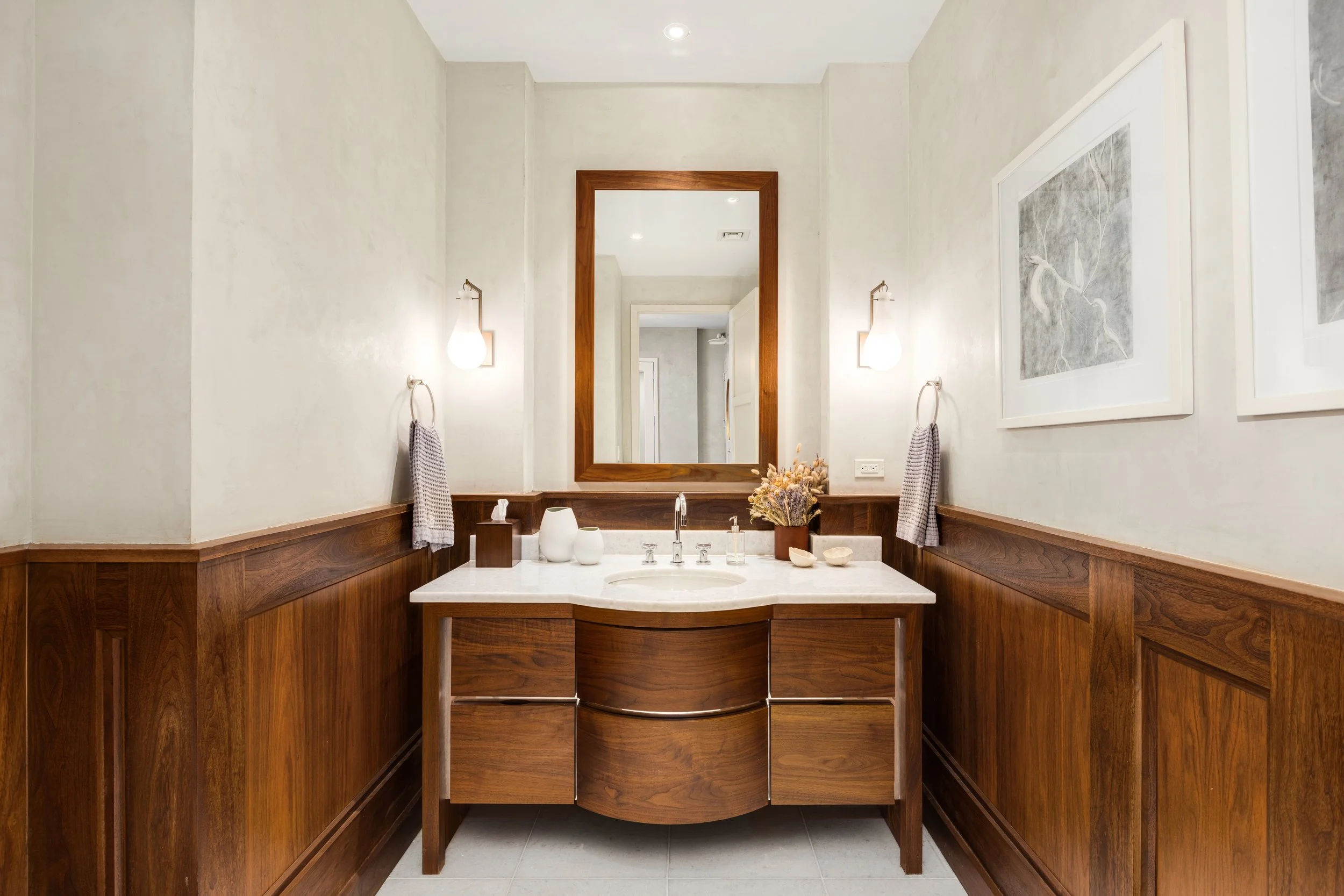 A bathroom vanity area with a wooden cabinet, a white marble countertop, a large framed mirror, and two wall-mounted lights.