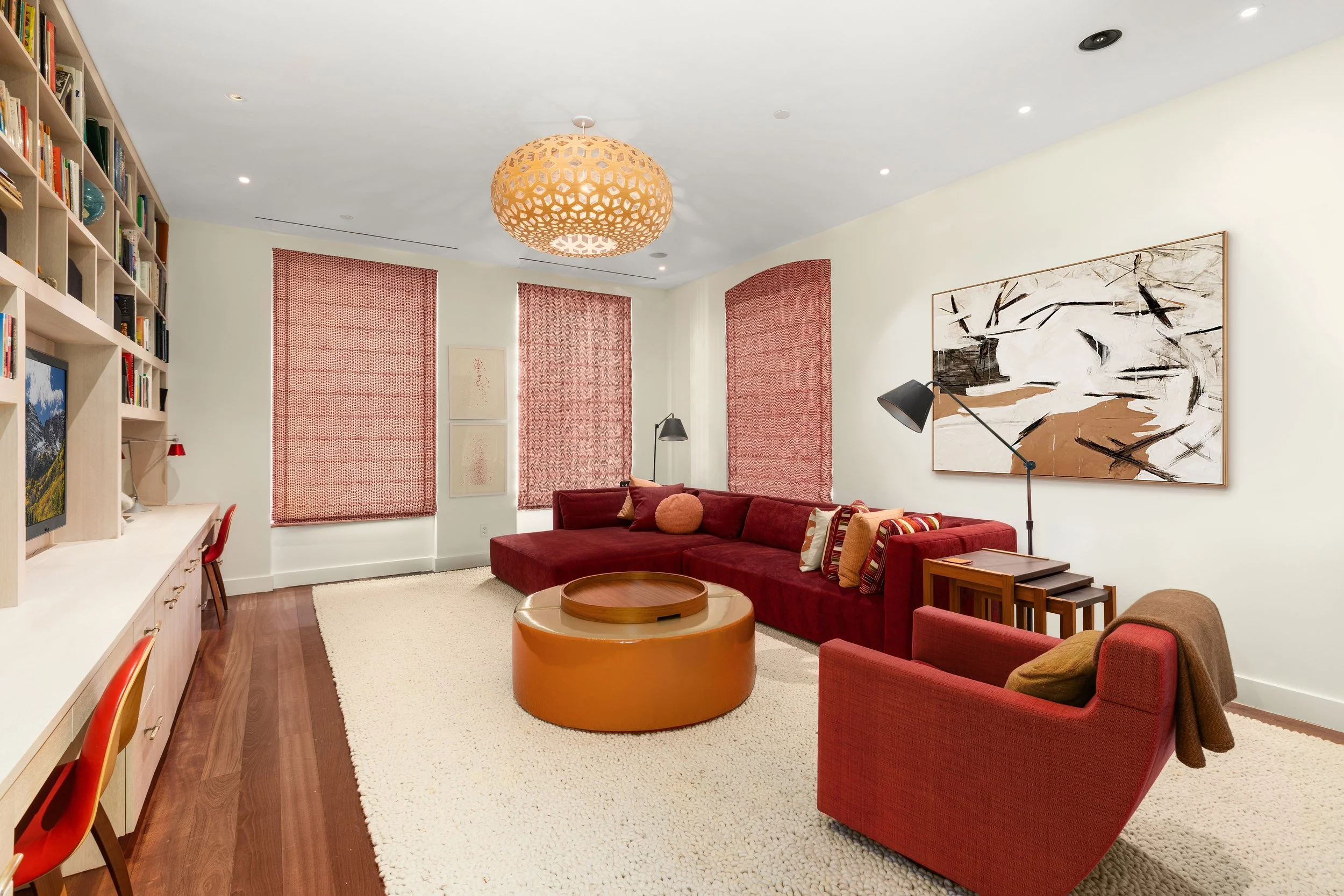 A living room with a large burgundy sectional sofa, red armchair, and wooden coffee table. There are bookshelves with a computer and decor, three windows with red shades, a large abstract painting on the wall, and a modern ceiling light.