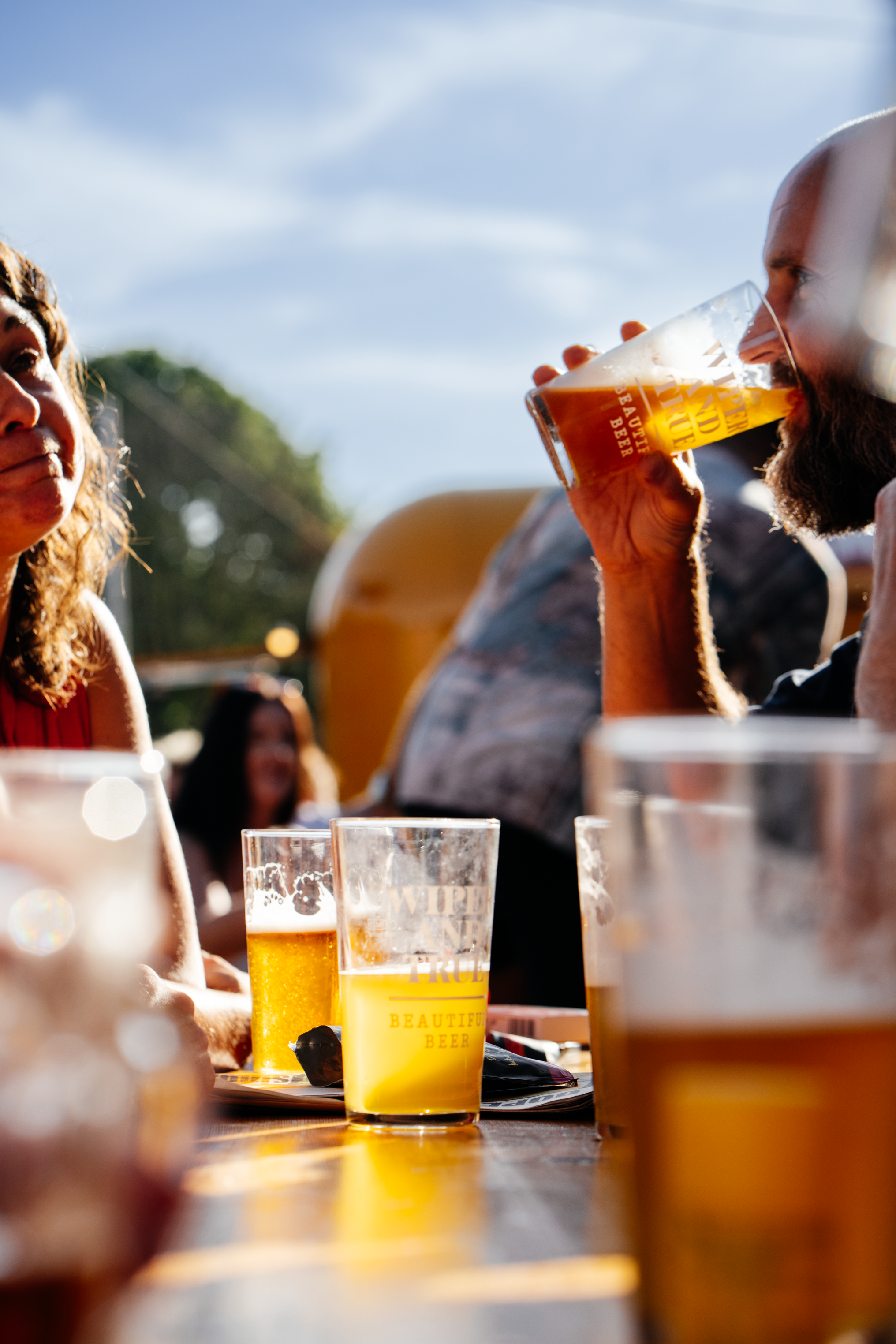 A man with a beard drinking beer from a Wiper & True glass at an outdoor setting with a woman nearby, in sunlight.