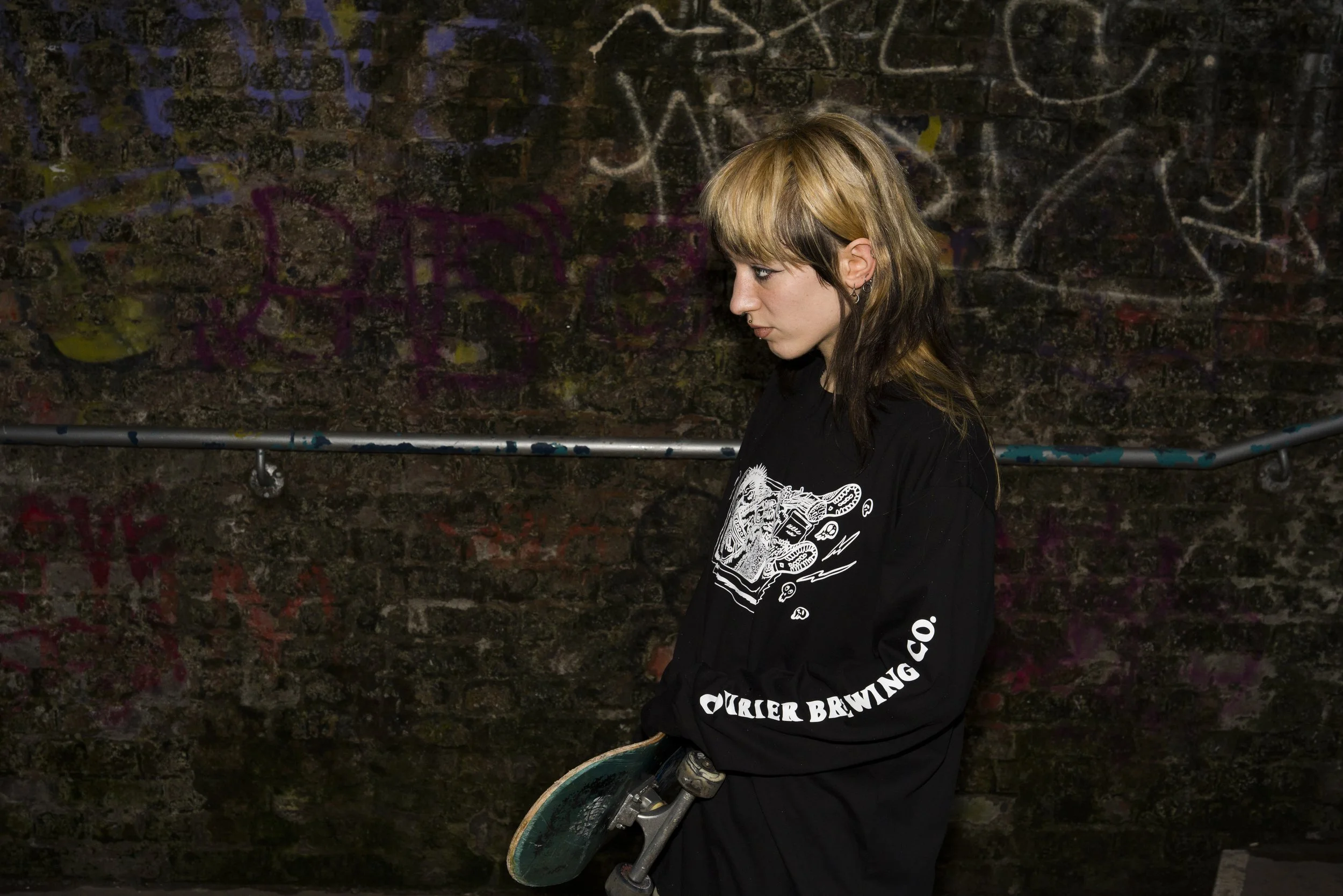 A young person with blonde and brown hair and multiple earrings standing against a graffiti-covered brick wall, holding a skateboard.