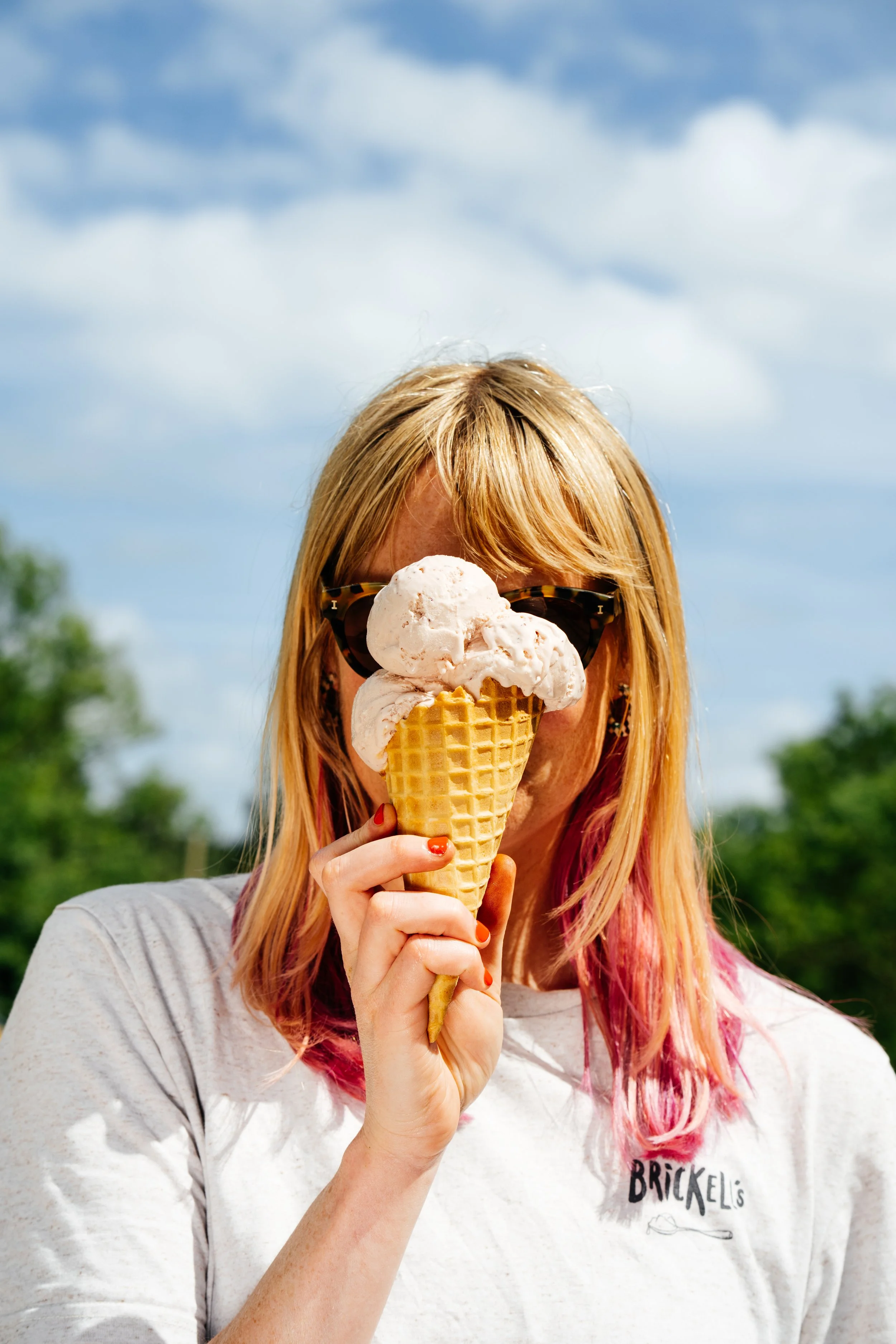 A woman with long blonde hair and colorful streaks holds an ice cream cone with three scoops in front of her face, obscuring it. She is wearing sunglasses and a gray t-shirt with the word 'BRICKELLS' printed on it. The background shows a partly cloudy sky and green trees.