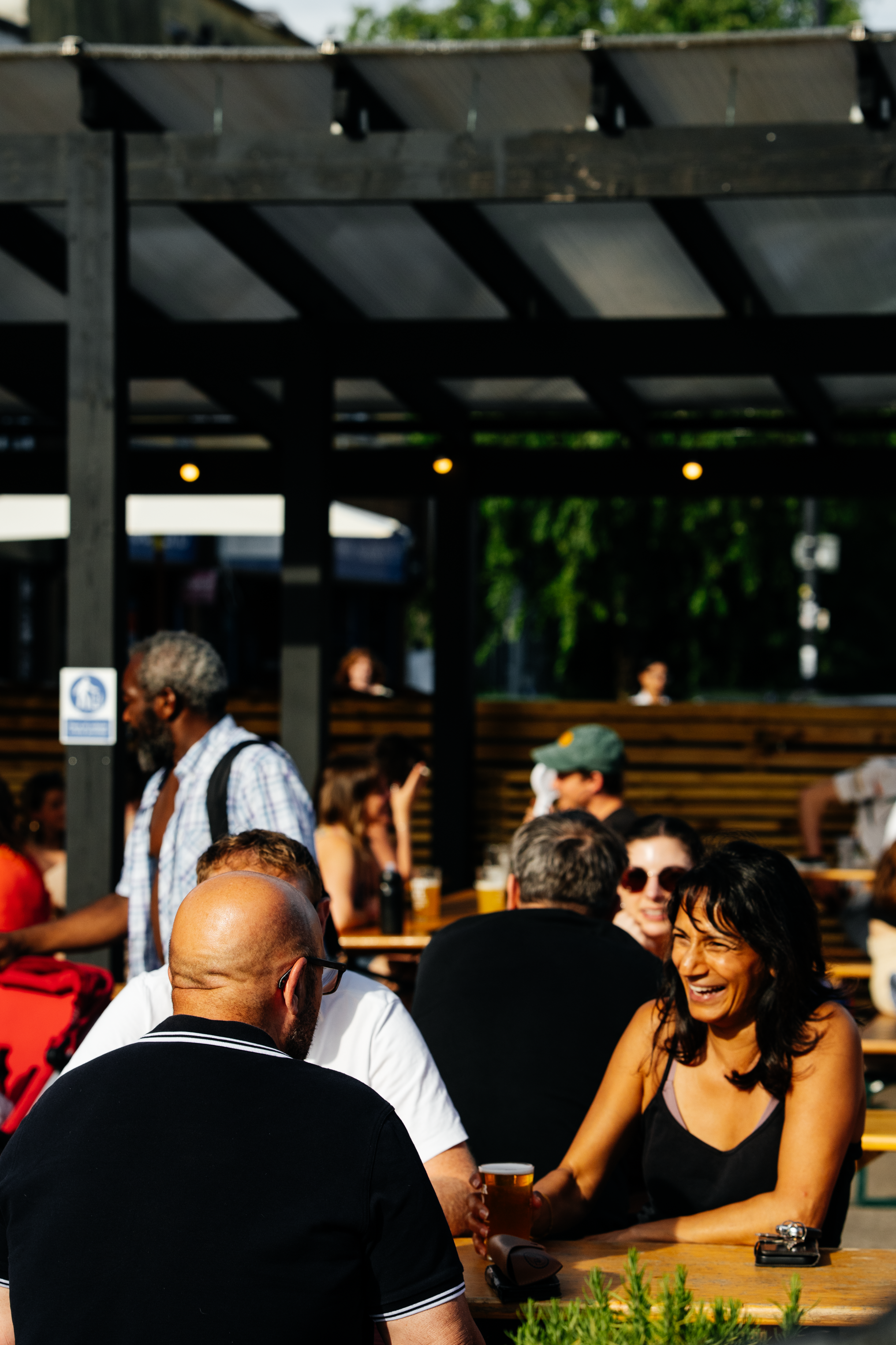 People socialising outdoors on a bench, some holding drinks, under a black pergola with string lights, in a lively setting with trees in the background.