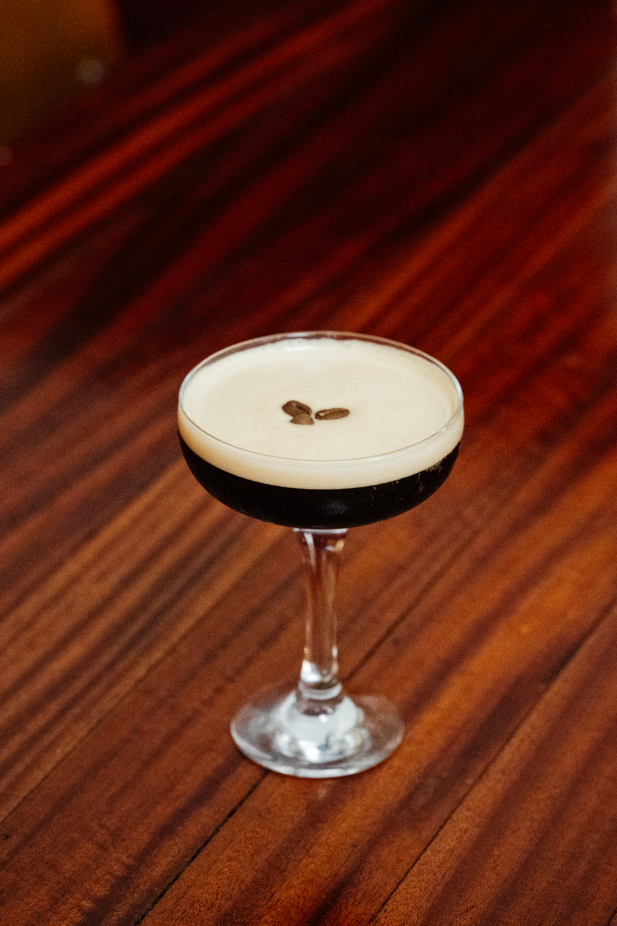 A cocktail glass with a dark beverage topped with a white layer and garnished with coffee beans, set on a wooden surface in Wiper & True Barrel Store, Bristol,