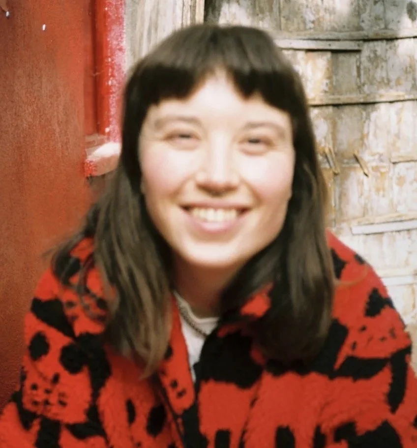 Nicci Peet smiling, wearing a red and black patterned jacket, sitting in front of a rust-colored wall and weathered wooden boards.