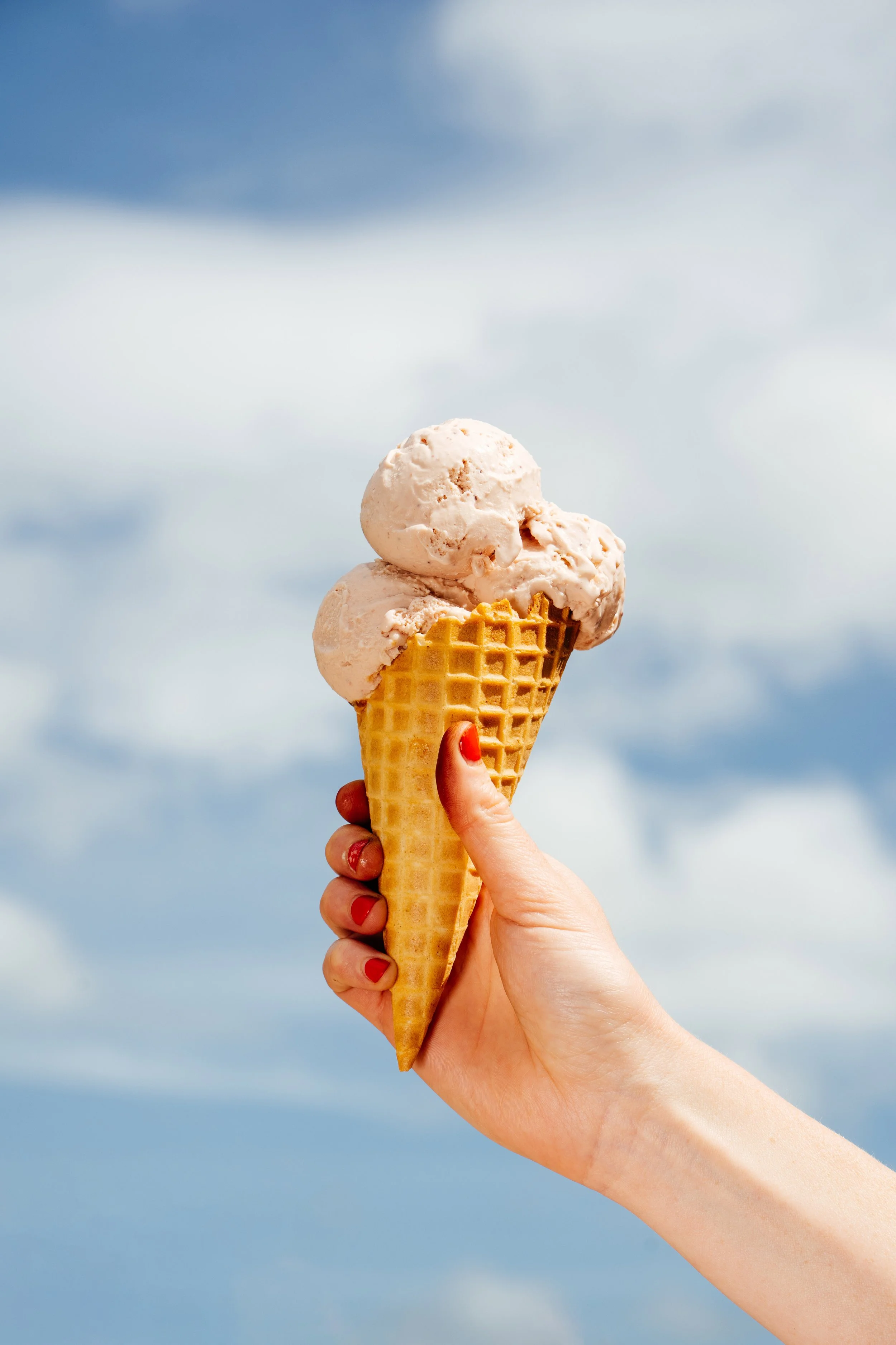 Hand holding an ice cream cone with three scoops of peach-colored ice cream against a blue sky with clouds.