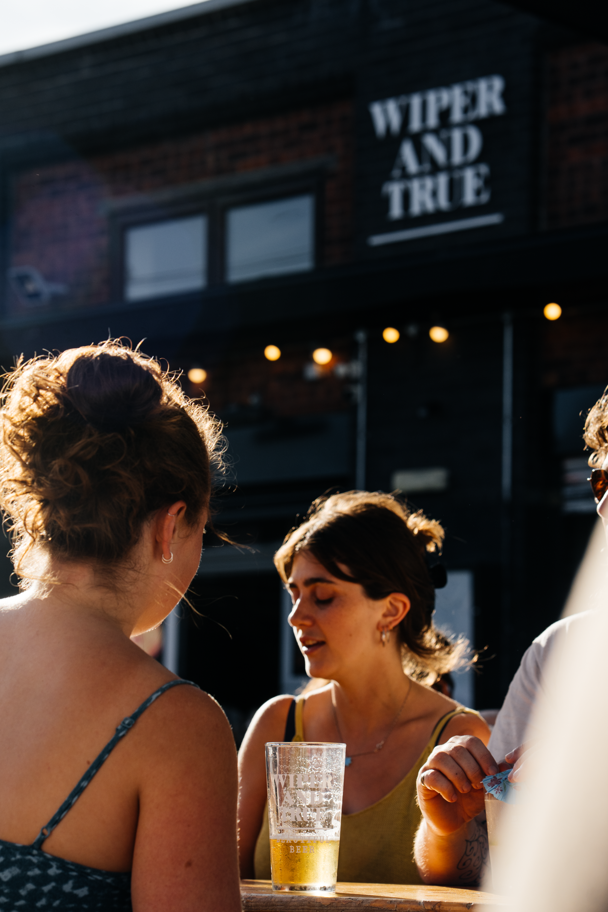 People socialising outdoors, with a man holding a pint of beer on a wooden surface, and a sign on the building that reads 'WIPER AND TRUE'.