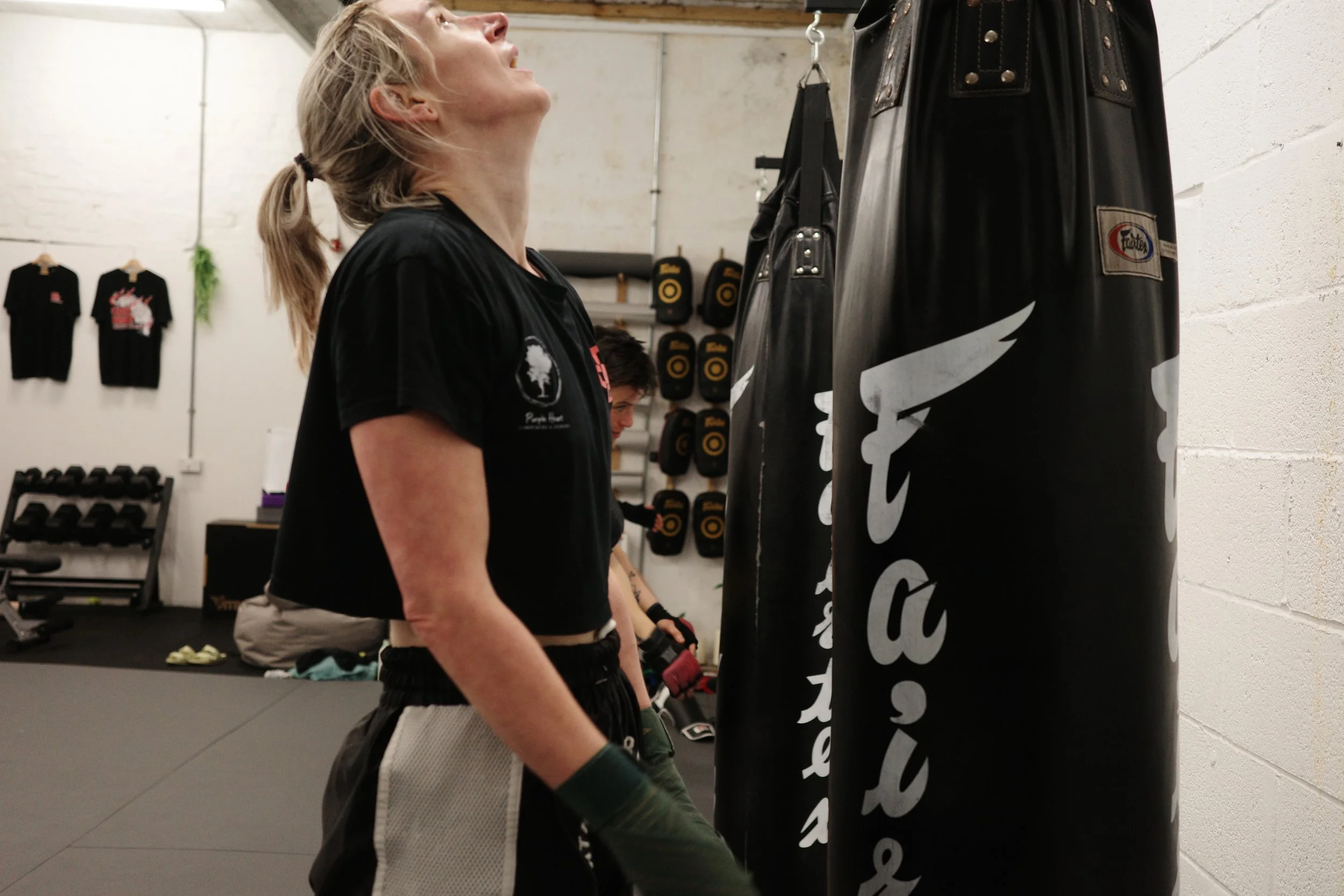 A woman with blonde hair tied back, wearing a black t-shirt and gloves, punches a black punching bag in a gym.