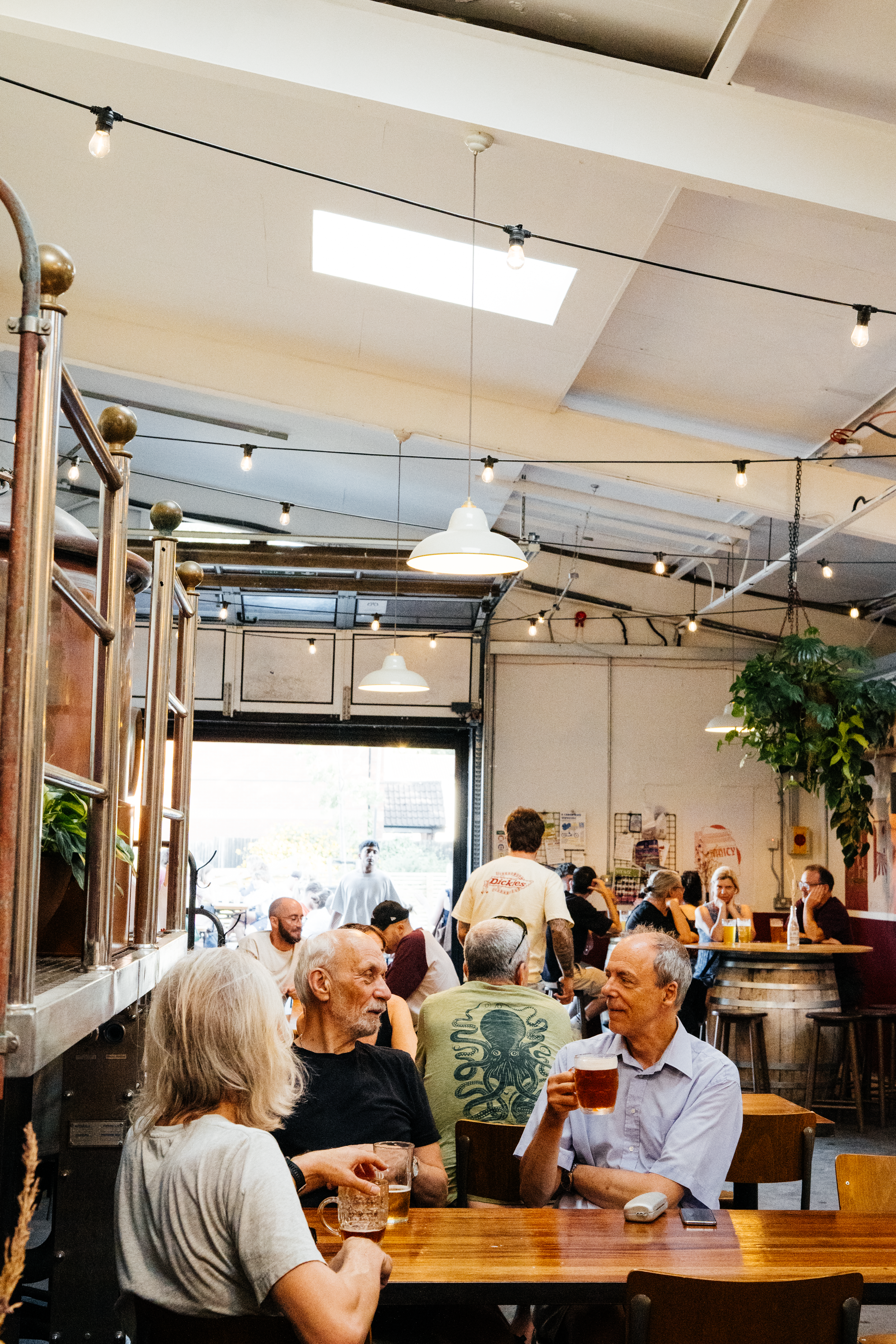 People are sitting at tables inside Wiper & True Barrel Store with string lights hanging from the ceiling. Large windows let in natural light, and one person is holding a glass of beer.