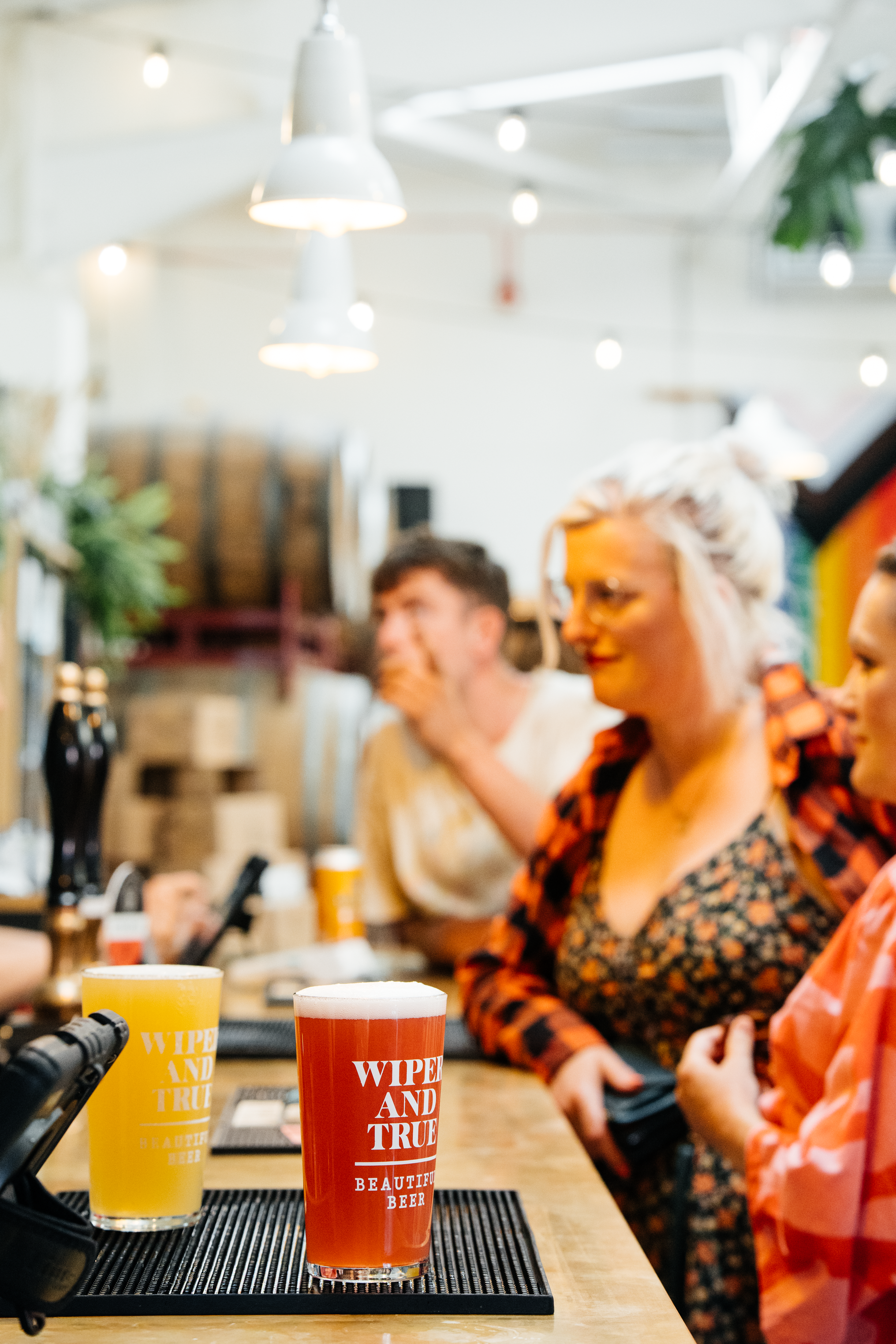 People sitting at the bar in Wiper & True Barrel Store with colourful drinks, including yellow and red beers, in a lively indoor setting with hanging lights and plants.