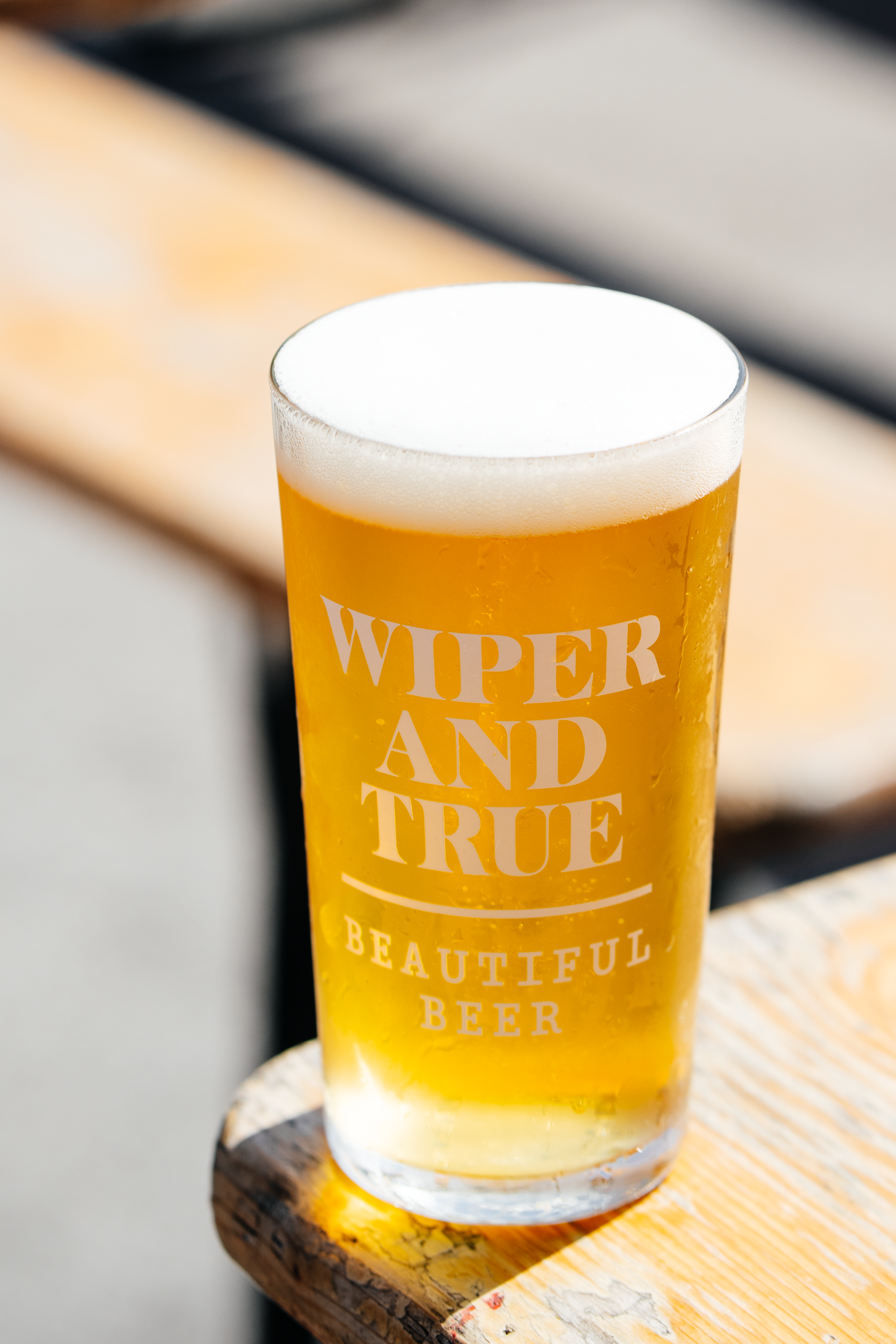 A tall glass of beer with a white head on a wooden table, with text on the glass reading 'WIFER AND TRUE BEAUTIFUL BEER'.