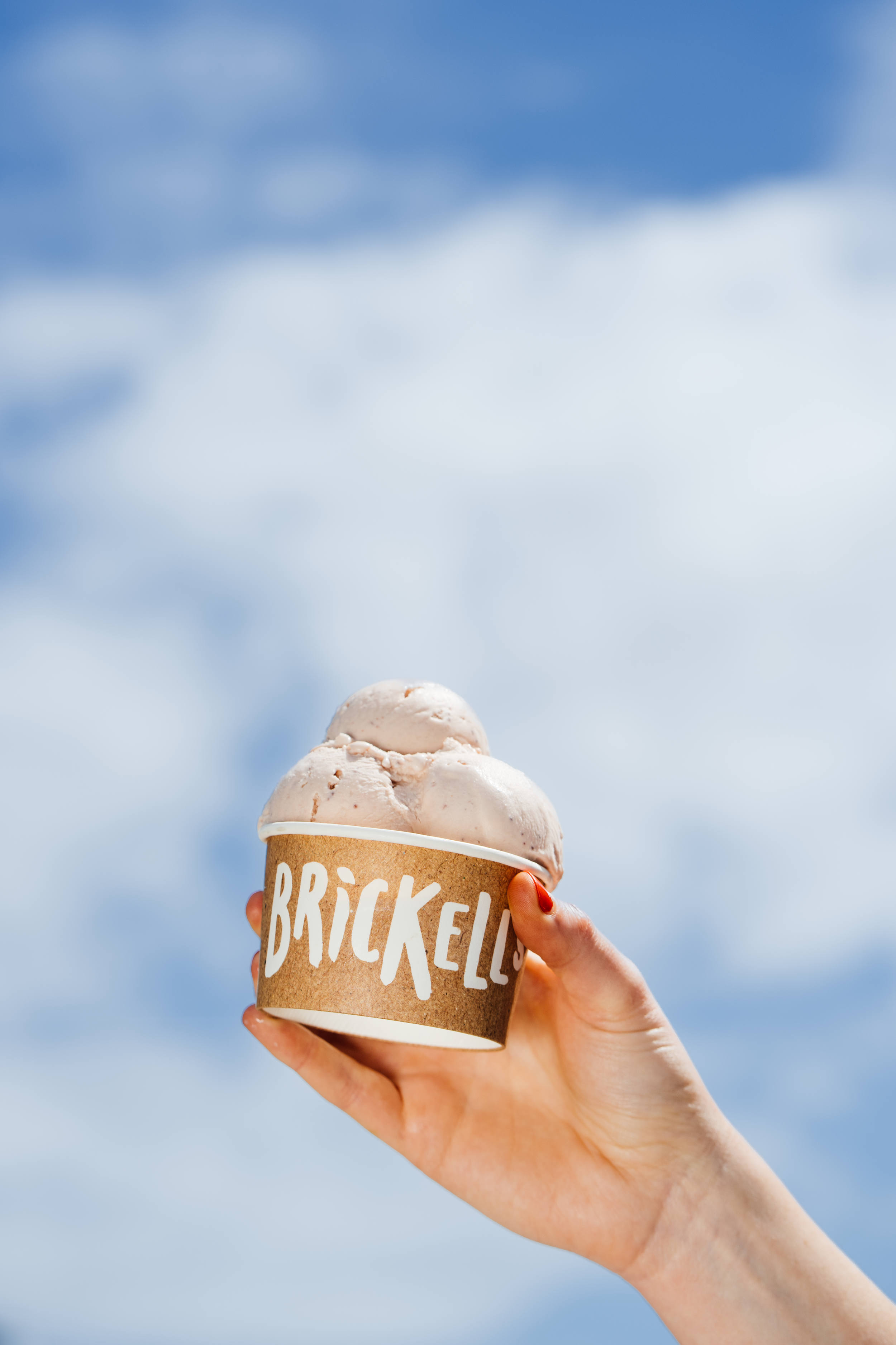 A hand holding a cup of ice cream with three scoops in front of a cloudy sky.