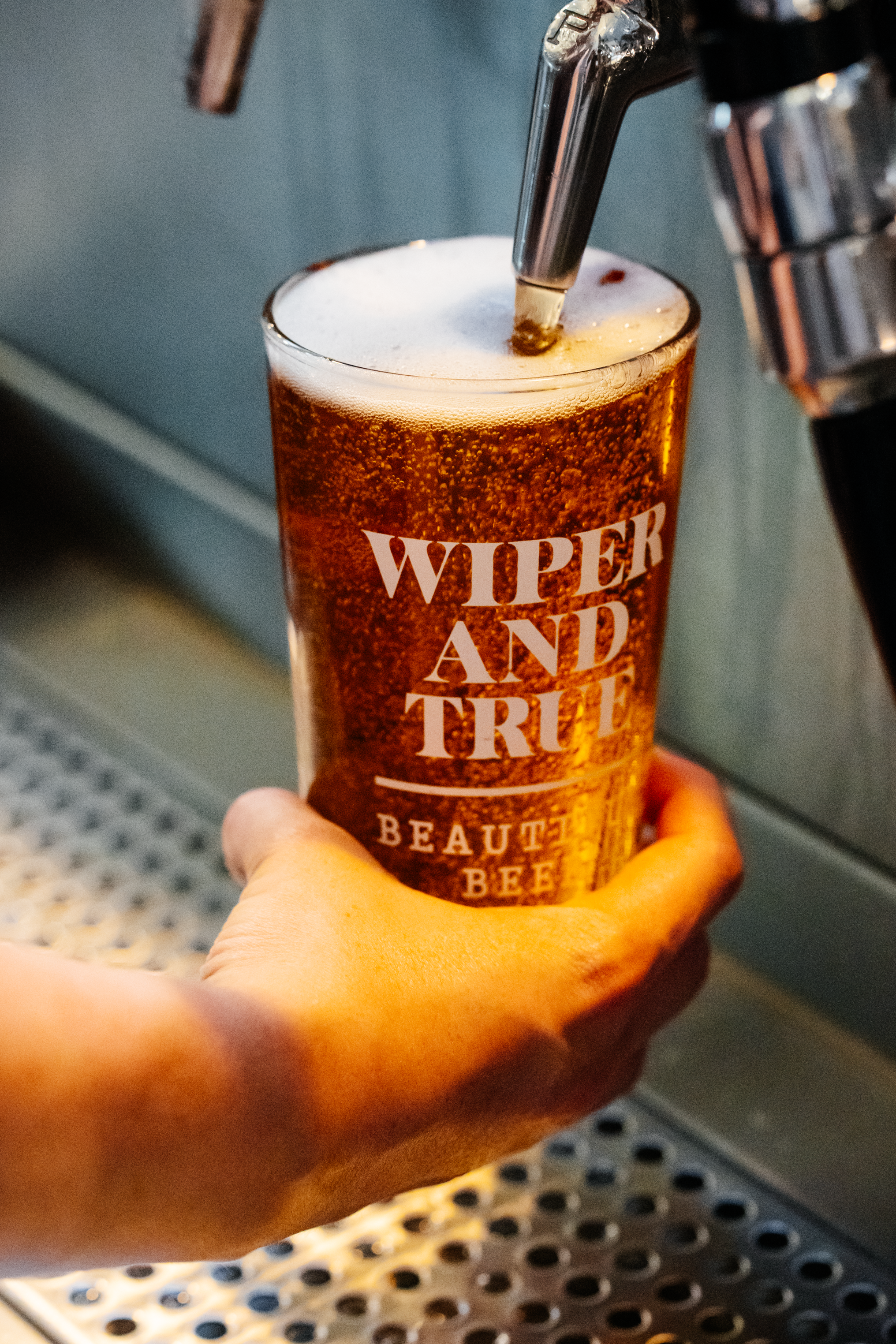A person's hand holding a pint glass filled with beer under a tap pouring beer into it, with the cup showing the text "Wiper and True" and a background of a metallic surface and a perforated tray.