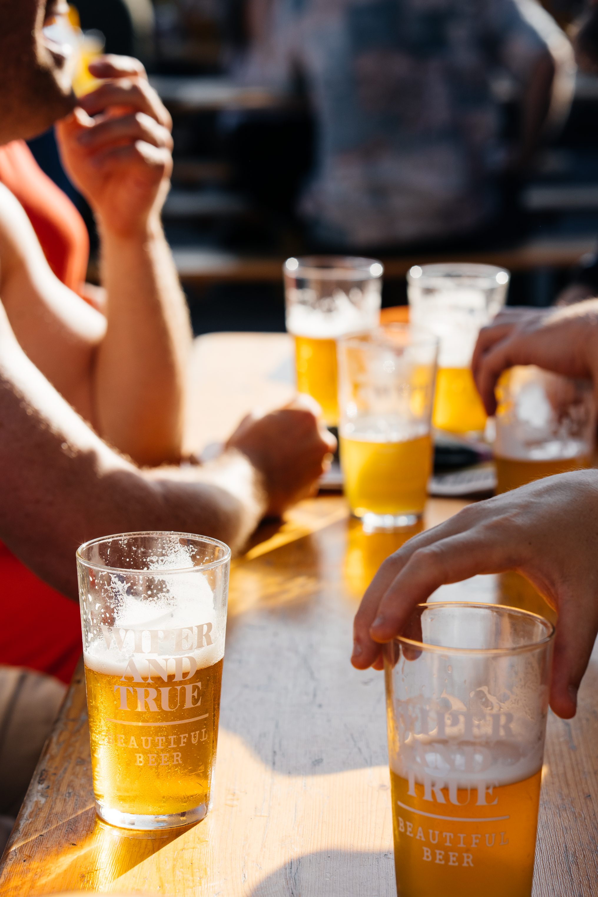 Close-up of several Wiper & True glasses of beer with foam on a wooden table at an outdoor setting, with people in the background.