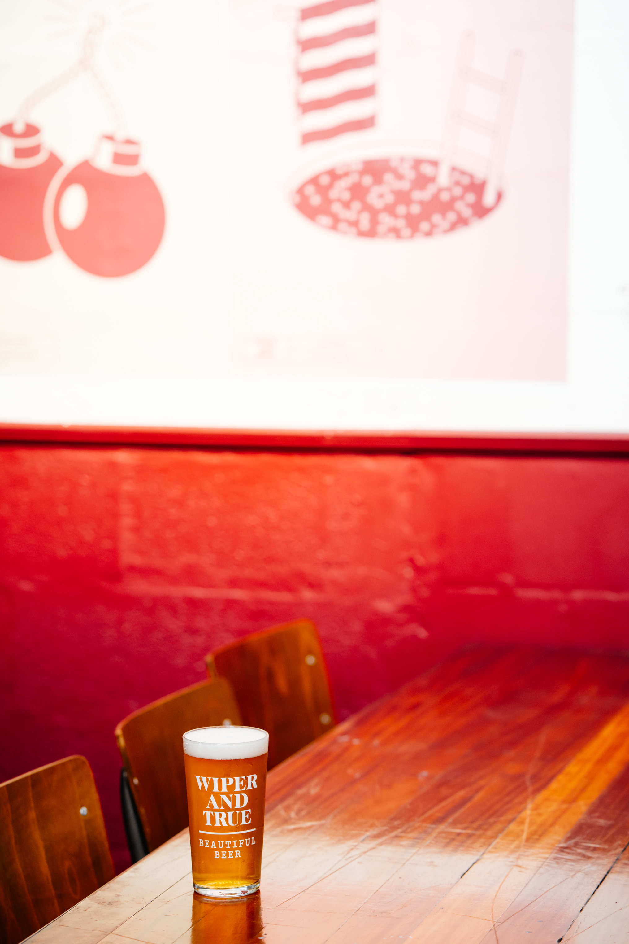 A pint glass of beer with the words "Wiper and True" and "Beautiful Beer" on it, sits on a wooden table in a bar or restaurant with pink walls and wooden chairs in the background.