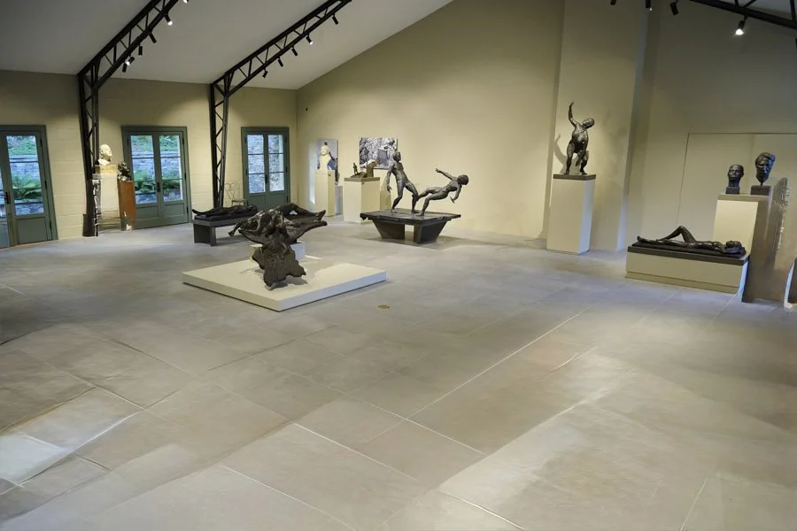 Art gallery with sculptures and busts displayed on pedestals and walls, featuring mythical creatures and human figures, with large windows and skylights letting in natural light.