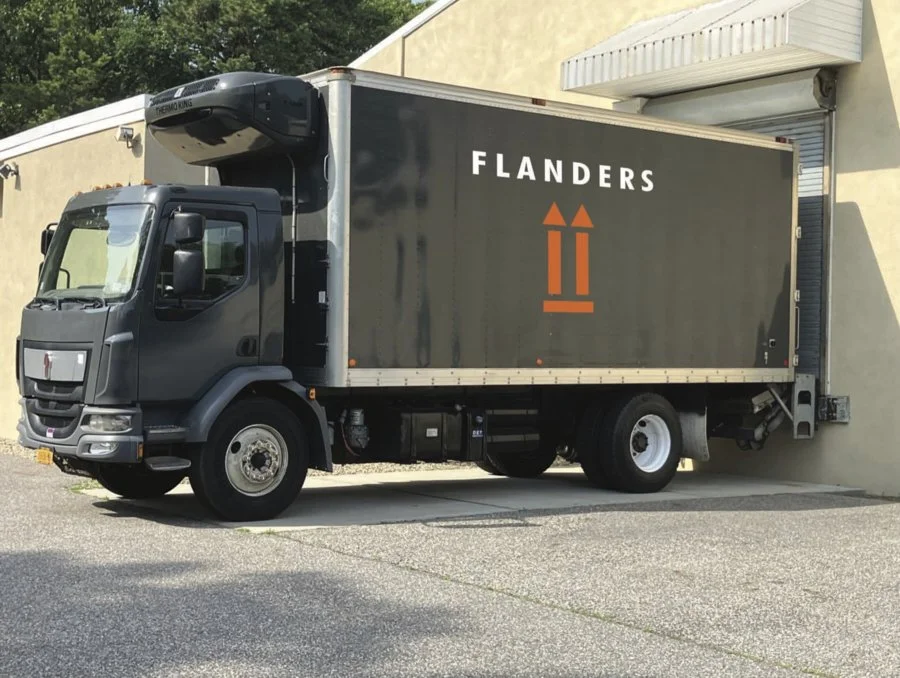 A black delivery truck with the word 'FLANDERS' on the side is parked next to a building with a roll-up door.