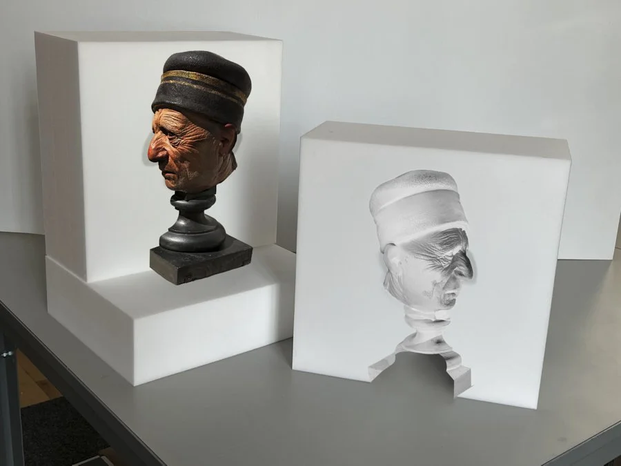 Bust of a man wearing a black cap with a gold band, split between a colorful sculpture and a white marble replica, displayed on white pedestals.