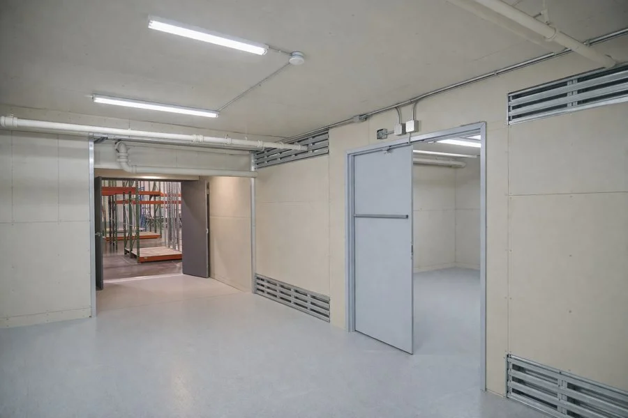 Industrial or storage space with plain walls, fluorescent lighting, metal door, and a view into a room with construction materials and scaffolding.