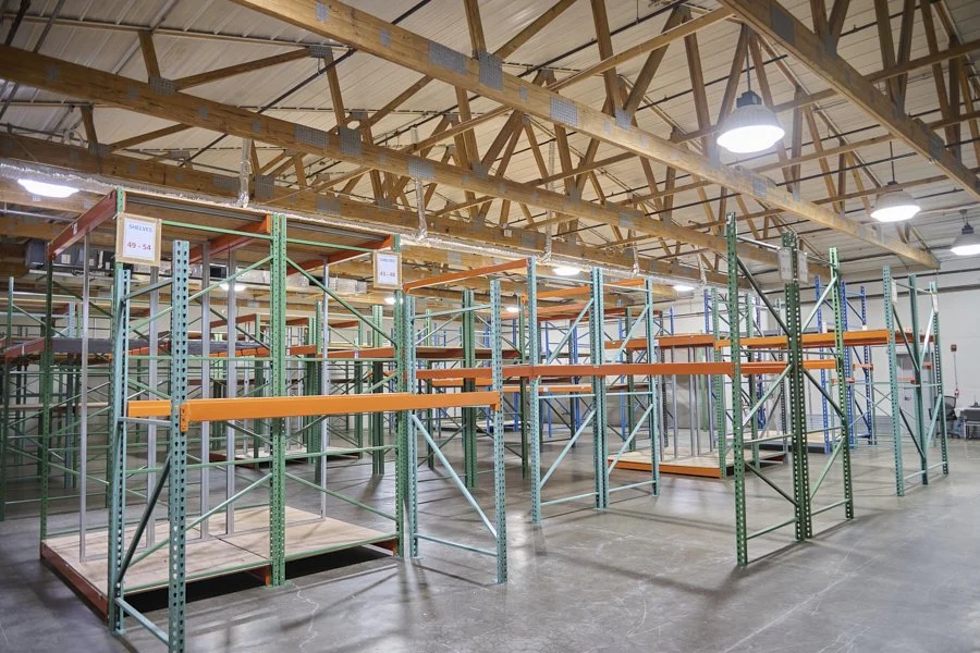 Empty warehouse with tall metal shelving units and orange beams, under a ceiling with exposed wooden beams and hanging industrial lights.