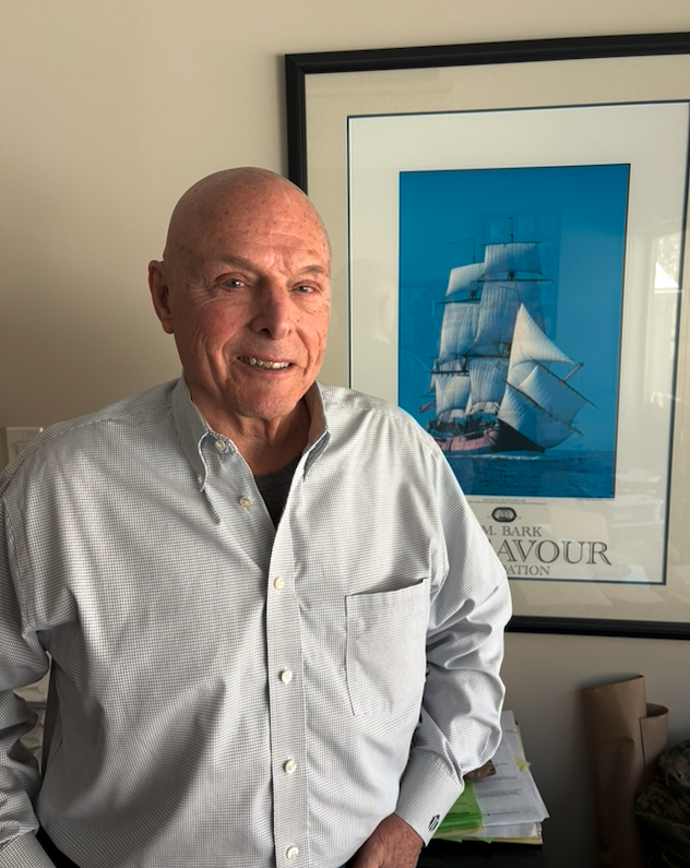 An elderly man with a bald head wearing a light-colored checkered button-up shirt, smiling and standing indoors in front of a framed picture of a ship with white sails on a blue background.