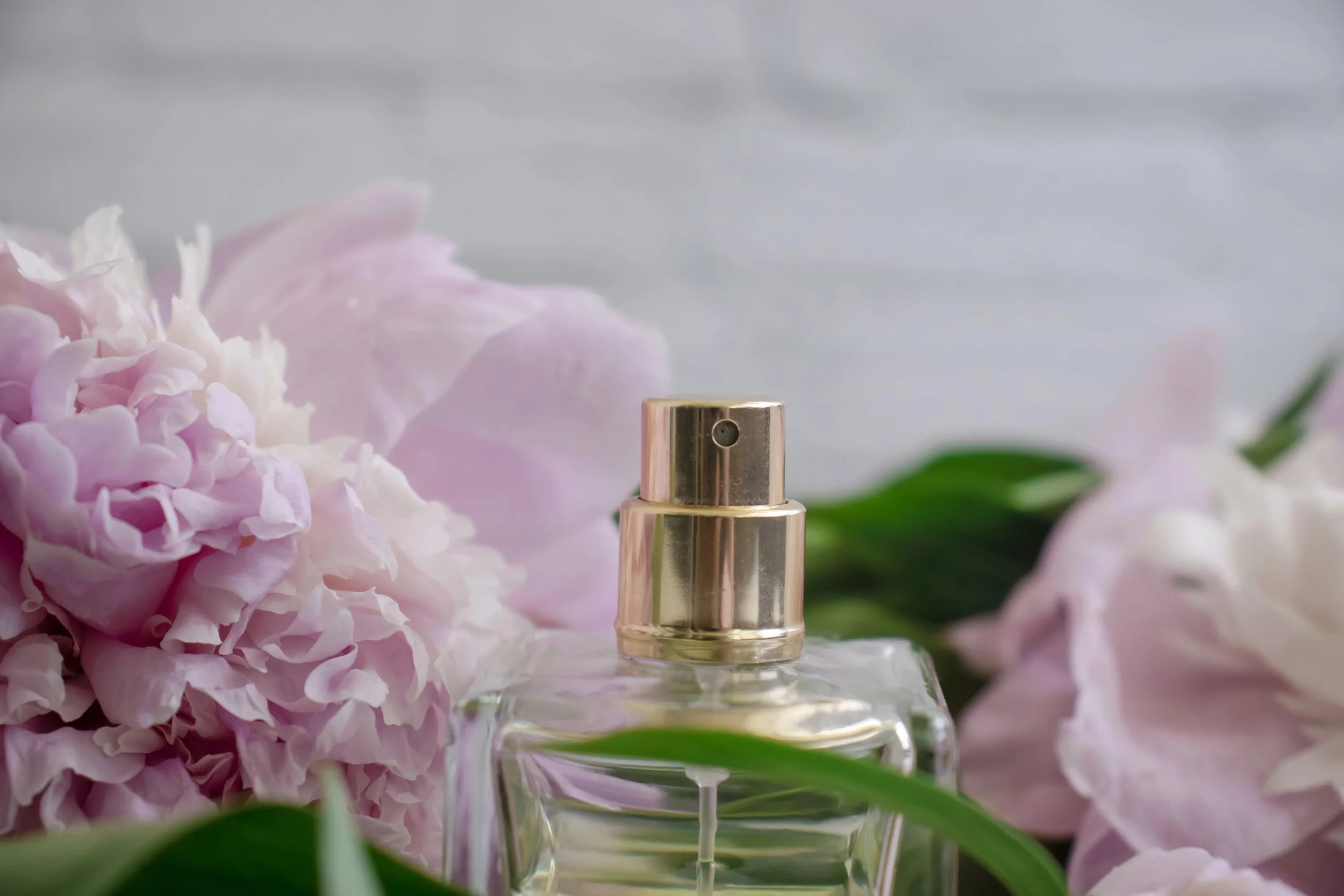 Frasco de perfume transparente con tapa dorada, rodeado de flores rosadas y verdes, sobre fondo gris borroneado.