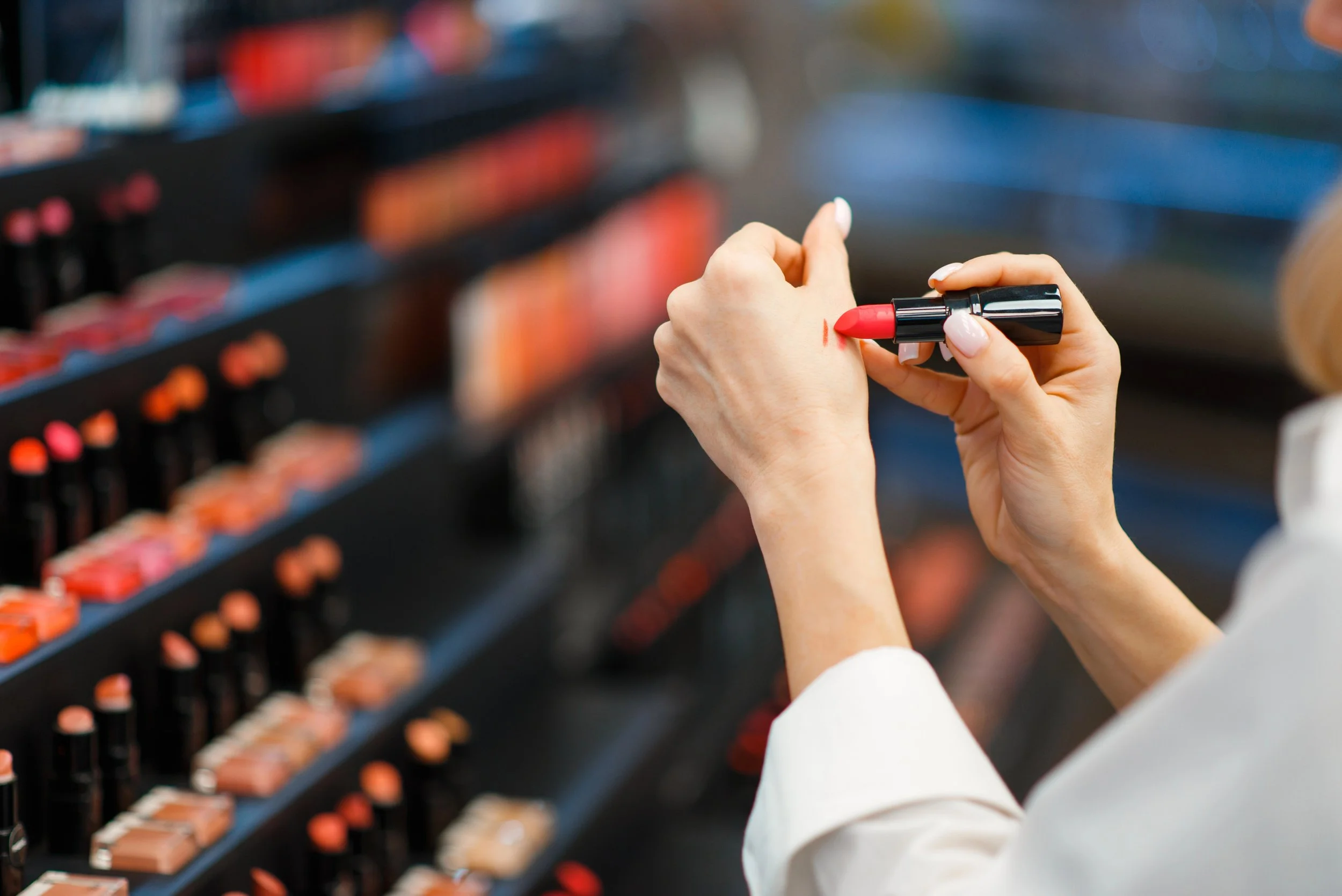Una persona sostiene un labial rojo y está aplicándolo en la mano, en una tienda con estantes llenos de productos similares y empaques en colores pastel y neutros.