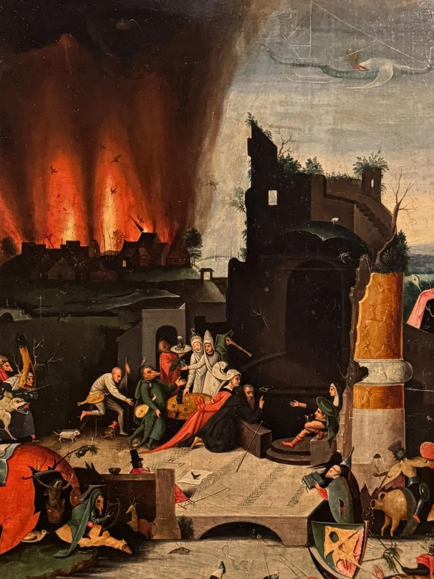 Detail of a Northern Renaissance painting showing a burning town under a dark plume of smoke, with small figures gathered around a well and scattered scenes of chaos and punishment in the foreground.