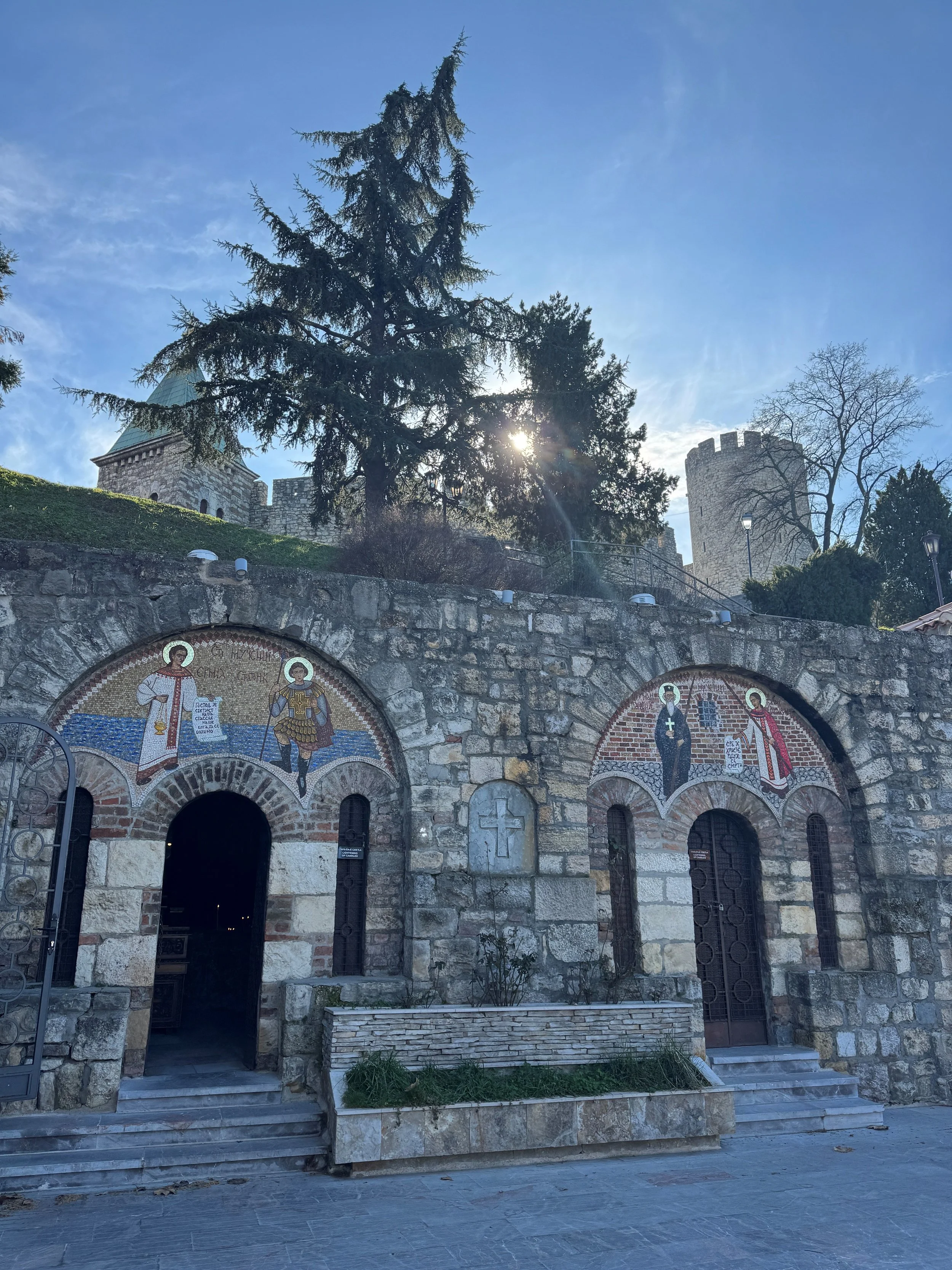 Exterior of the Chapel of Saint Petka in Belgrade, showing two arched entrances set into a stone wall, each topped with colourful mosaic saints, with trees and fortress towers rising behind in bright daylight.
