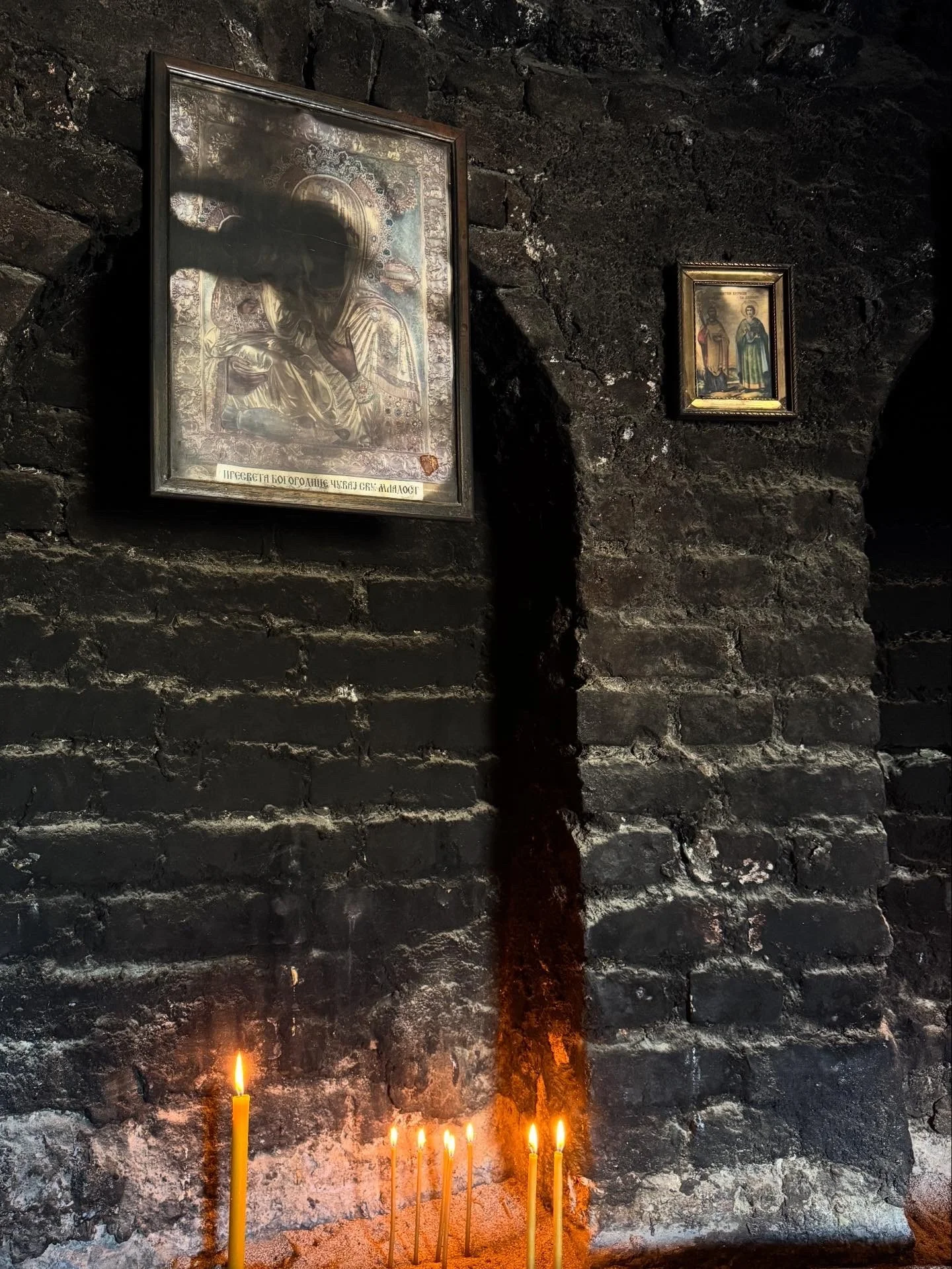 Soot-darkened stone wall inside the Chapel of Saint Petka in Belgrade, with framed icons above a row of lit candles whose smoke blackens the Virgin’s face.