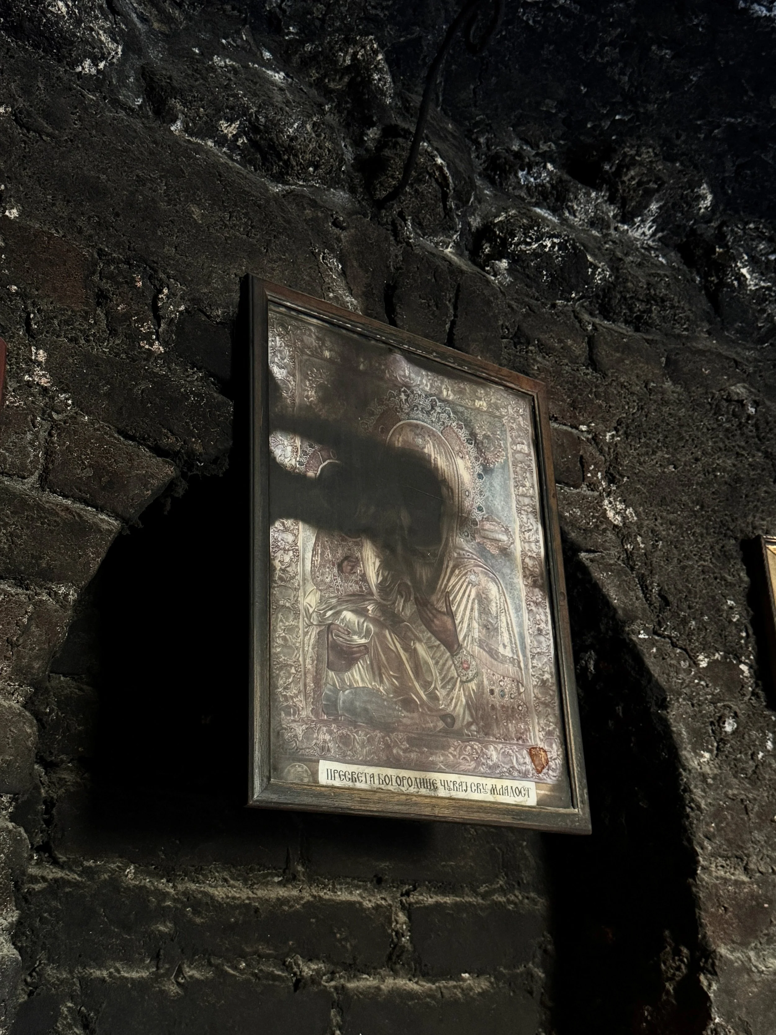 Framed icon of the Virgin and Child mounted on a soot-blackened stone wall inside the candle alcoves of the Chapel of Saint Petka in Belgrade, the saint’s face partially darkened by candle smoke.
