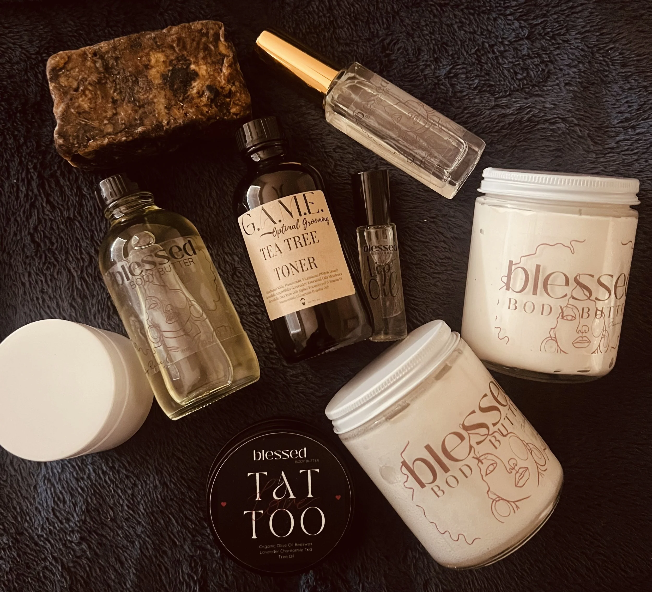 Collection of skincare and self-care products on a black fuzzy surface, including body butter, tea tree toner, body oil, lip oil, soap, and a container labeled 'Tat Too'.