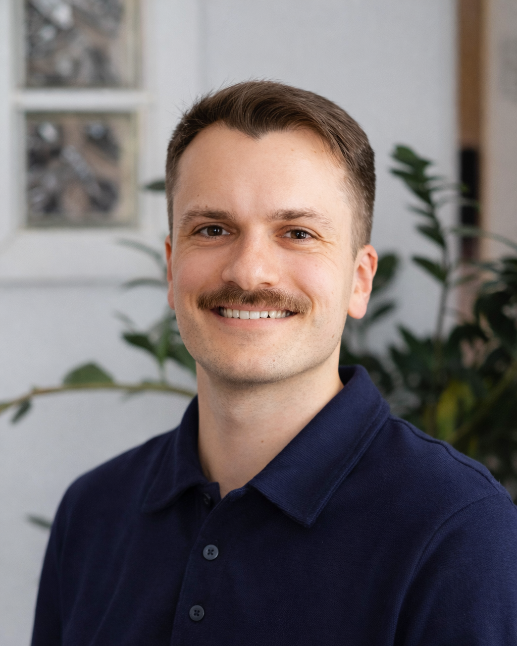 A young man with short brown hair, a mustache, and a smile, wearing a navy polo shirt, standing indoors near a plant and a window.
