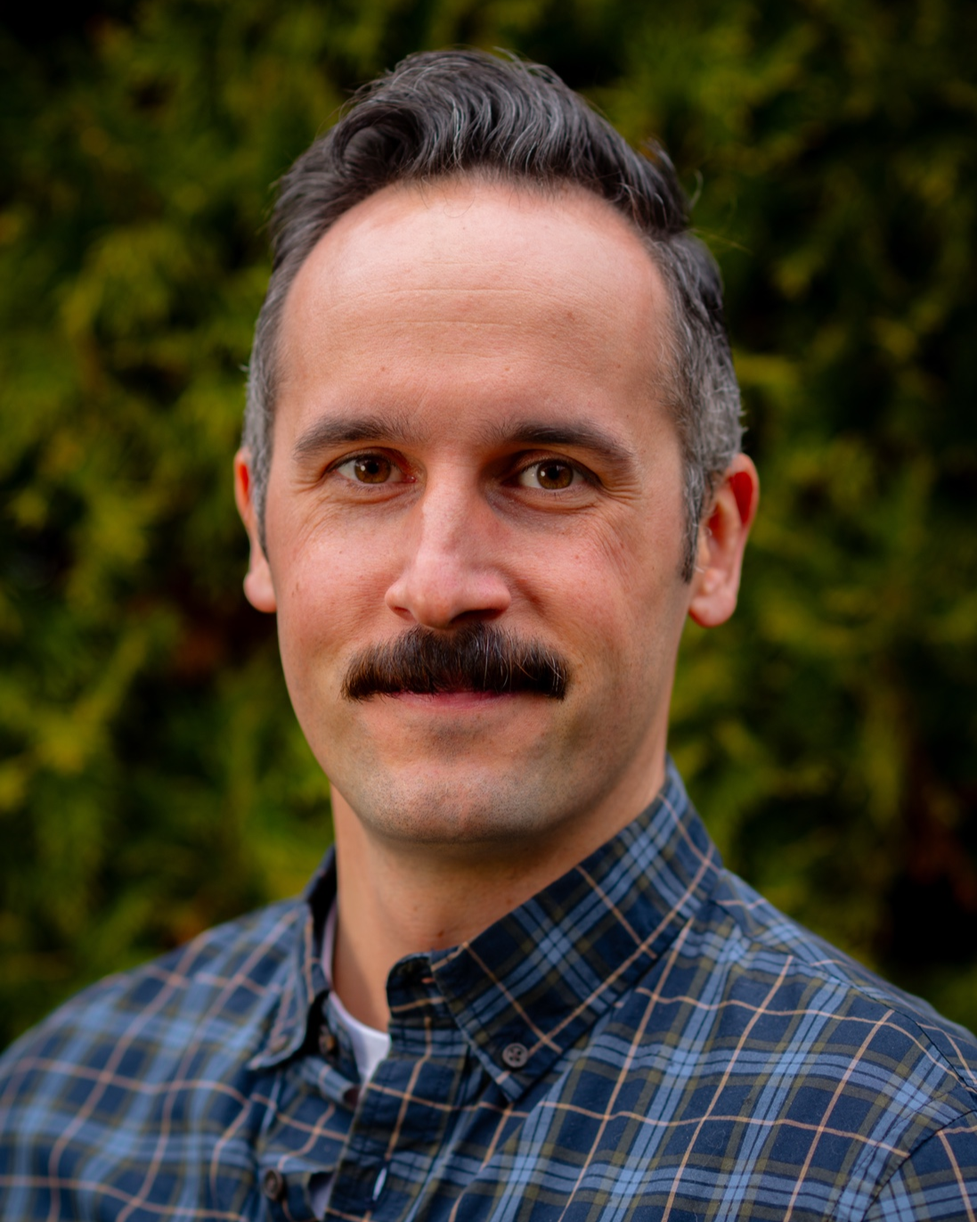 A man with dark hair, a mustache, wearing a plaid shirt, standing outdoors with green foliage in the background.