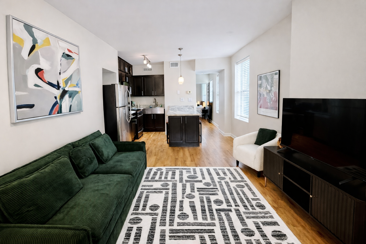 Living room with green sofa, black TV stand, white armchair, abstract wall art, black kitchen cabinets, illustrated artwork, hardwood floors, patterned rug, kitchen area with stainless steel appliances, small dining area in background.