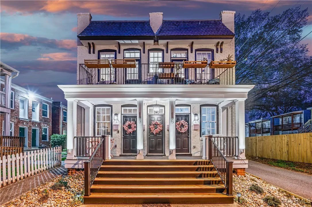 Three-story house with black front doors decorated with pink wreaths, white pillars, porch lighting, and wooden stairs, with a balcony and a night sky in the background.