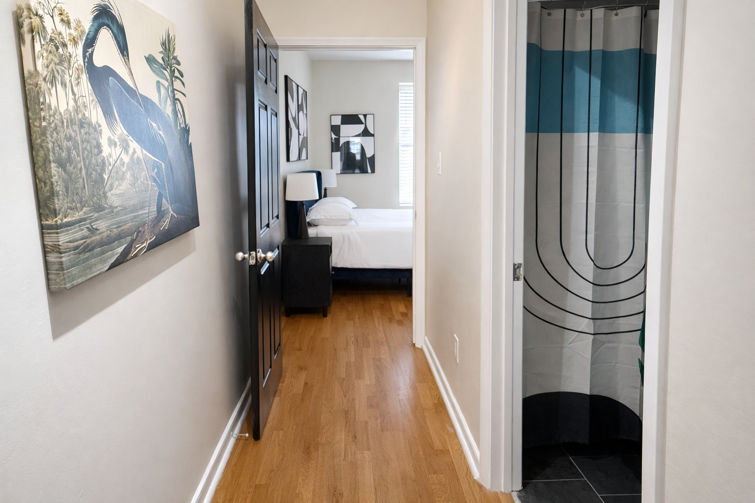 Hallway view towards a bedroom with an open door, visible bed with white bedding and pillows, bedside table with lamp, and wall art, with a bathroom shower stall on the right.