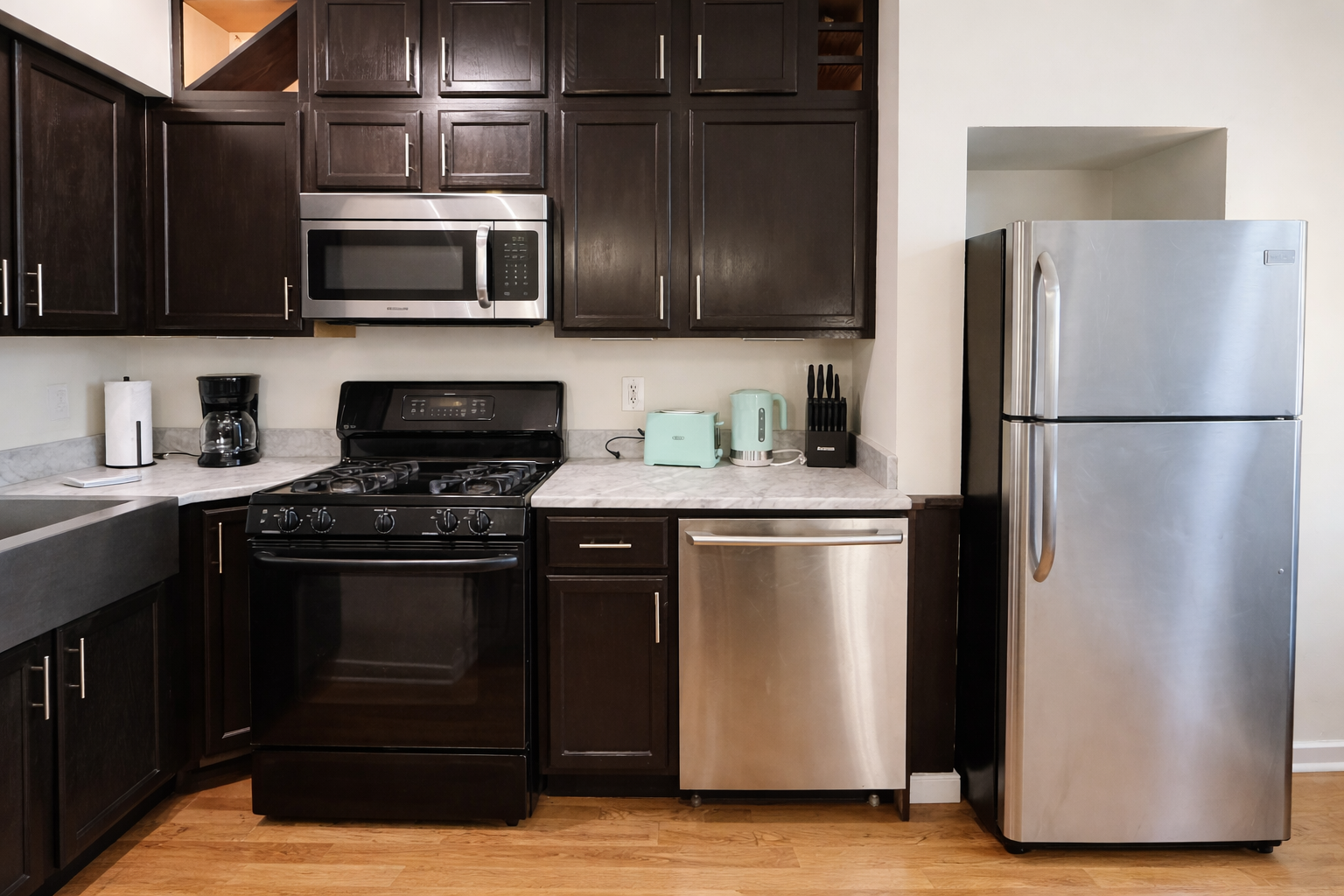 A modern kitchen with dark wood cabinets, a black gas oven, stainless steel microwave, and refrigerator. The countertop has small appliances including a coffee maker, toaster, and electric kettle.