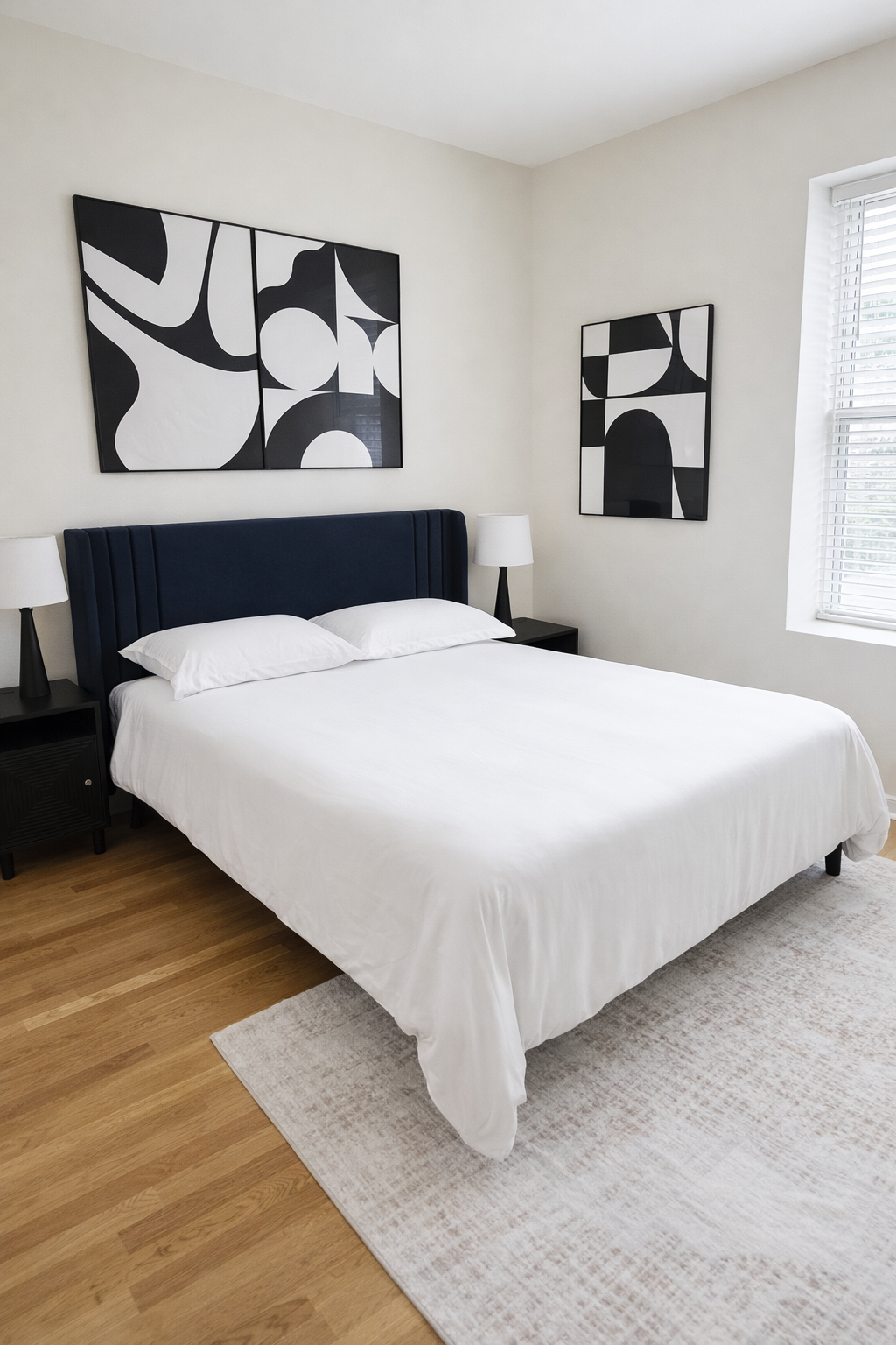 A modern bedroom with a white bed, navy blue upholstered headboard, two black bedside tables with white lamps, abstract black and white artwork on the wall, a window with white blinds, and light wood flooring with a beige area rug.