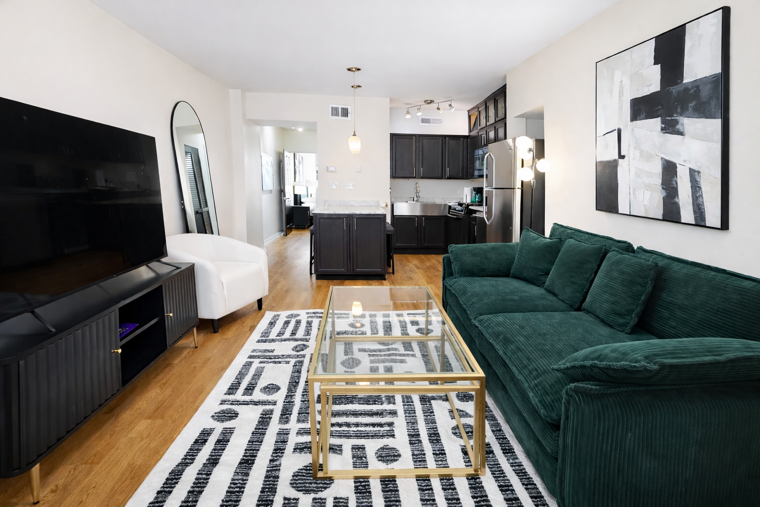 Living room with a dark green velvet sofa, a white armchair, a black TV stand with a large flat-screen TV, a black and white patterned rug, a glass-top gold coffee table, and artwork on the walls. The open kitchen with dark cabinets, a stainless stee