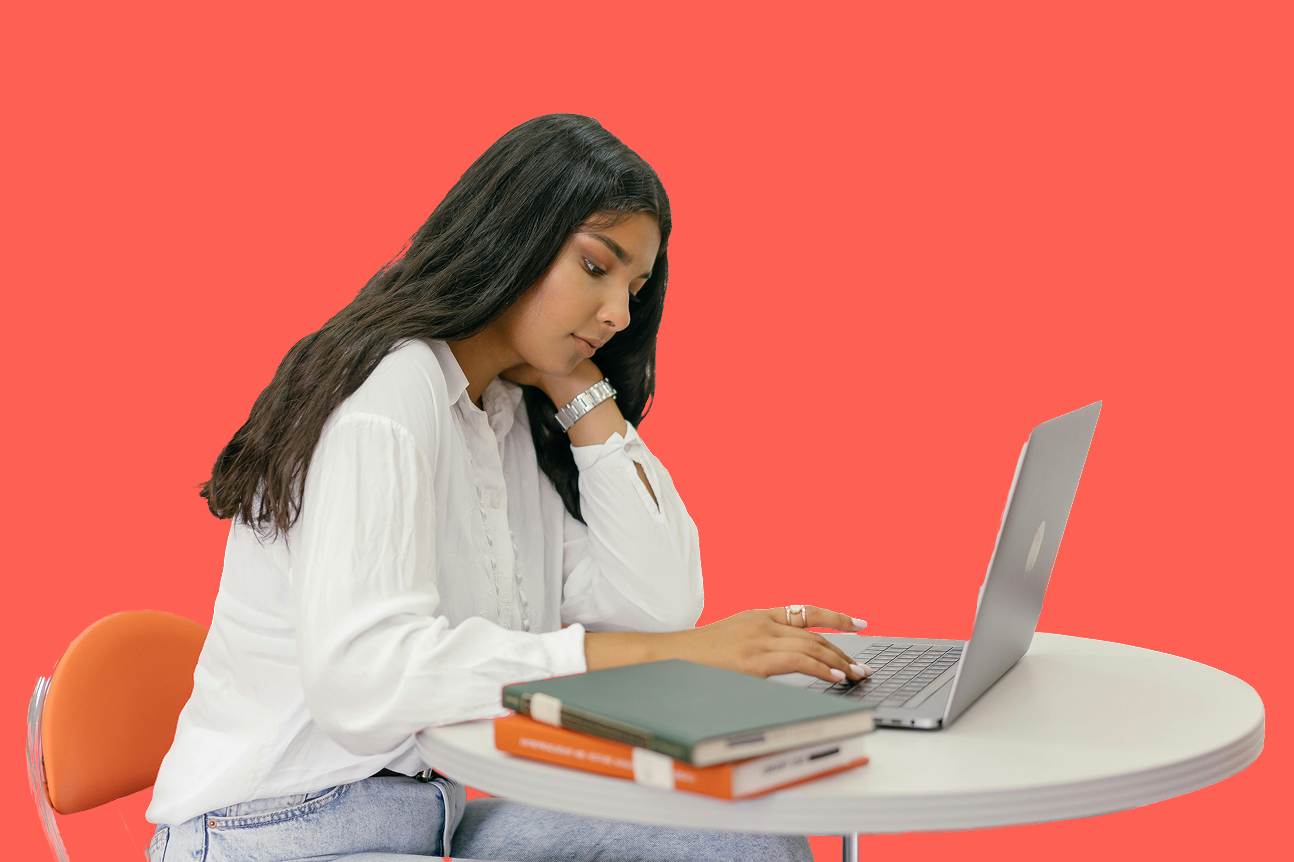 A young woman is sitting at a round white table with a coral background. She has long dark hair and is wearing a white shirt, a watch, and rings. She is using a silver laptop, with a few notebooks stacked on the table.