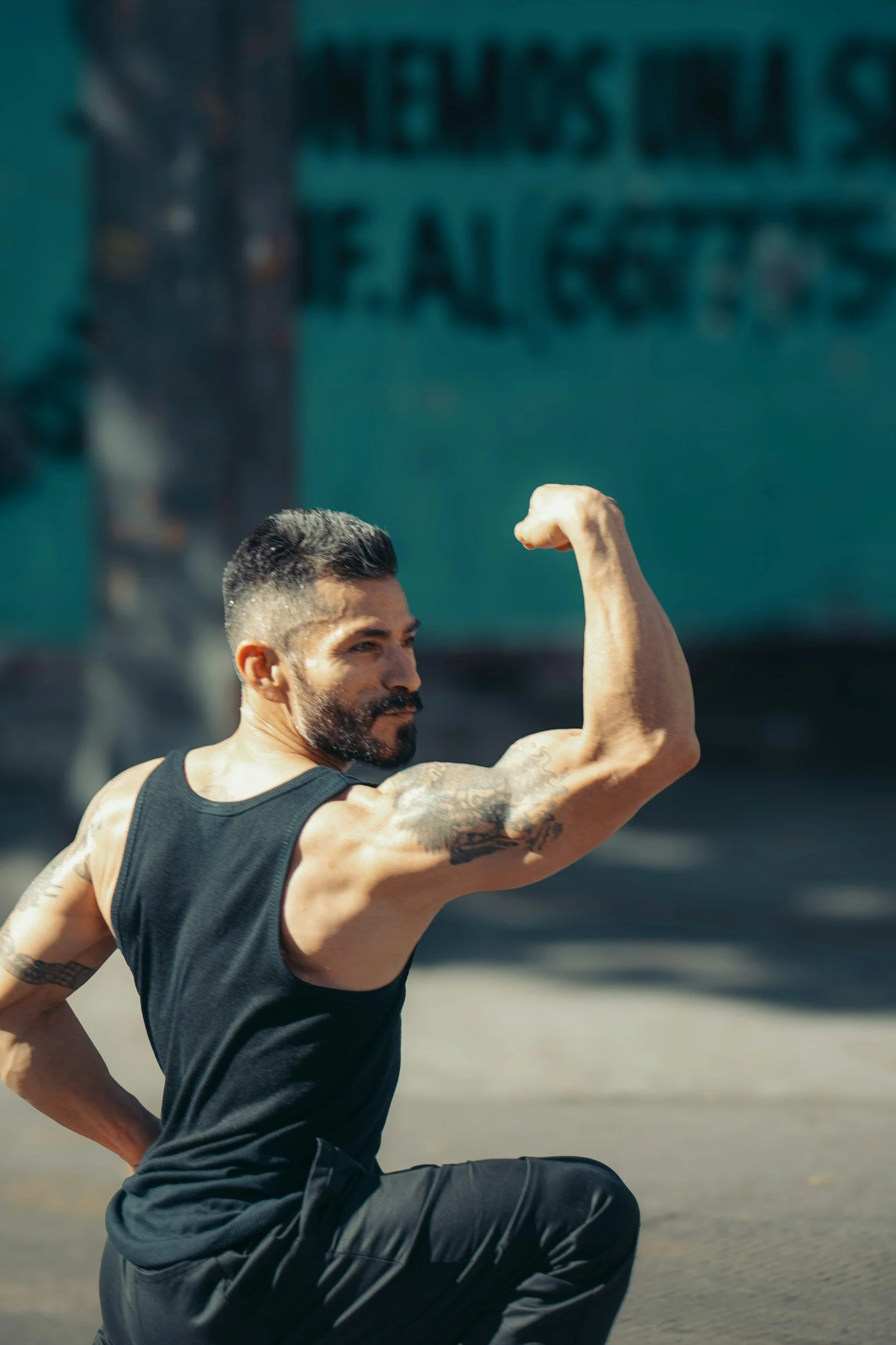 A man with dark hair and beard, wearing a black tank top, is outdoors during daytime. He is flexing his right arm, showing his bicep, with his left hand on his hip, against a background of a blue-green wall with text and a street sign.