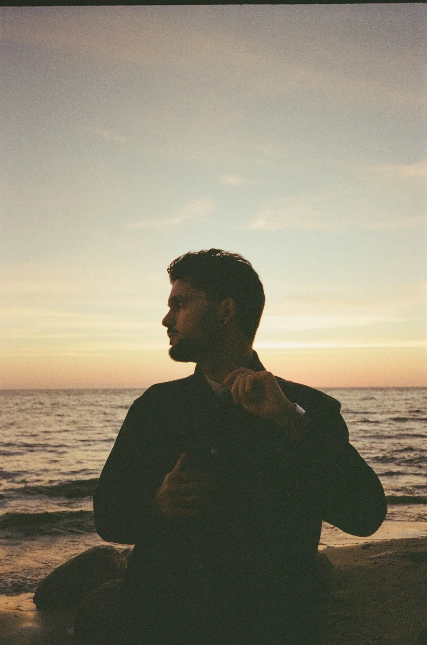 A silhouette of a young man with a beard at the beach during sunset, standing near rocks with the ocean in the background.