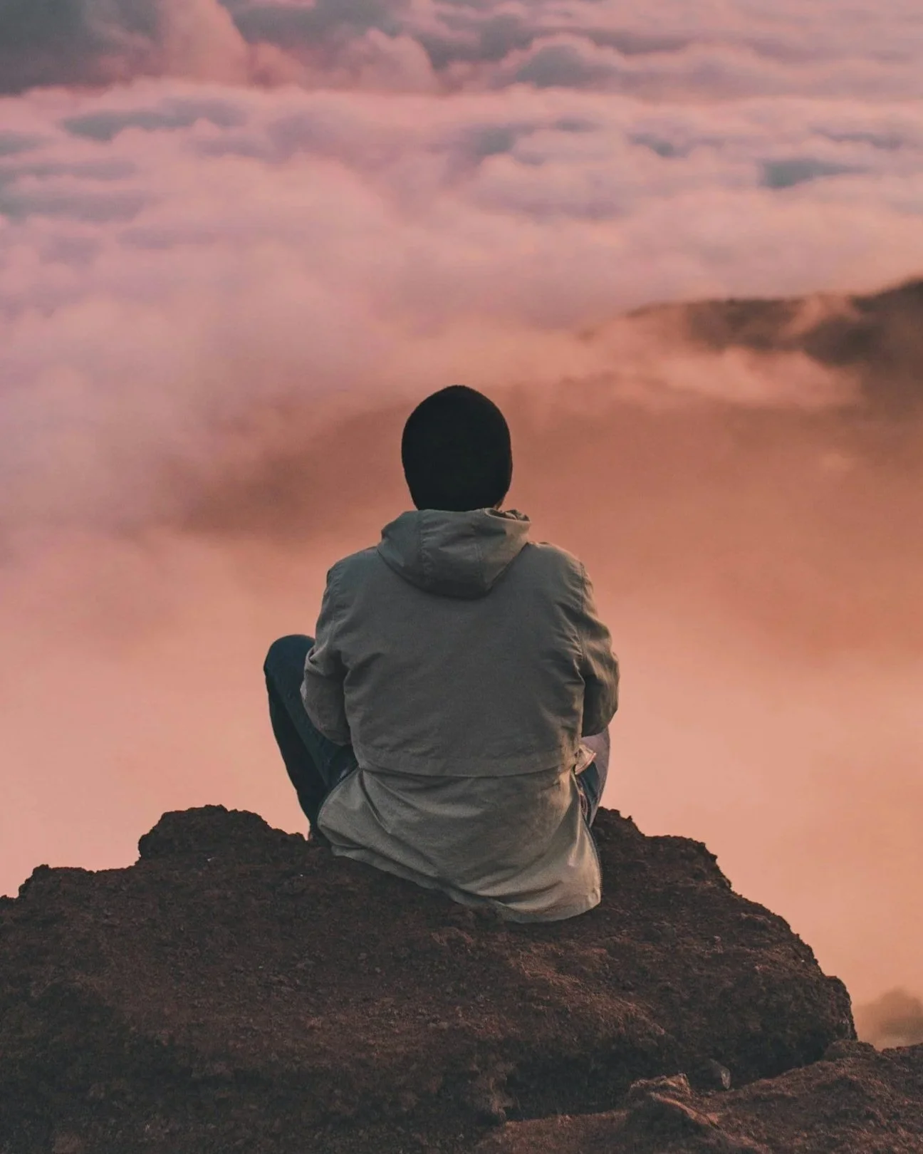 A person wearing a light-colored jacket and black pants sitting on a rocky surface, facing away, looking at a sky filled with pink and purple clouds at sunset or sunrise.