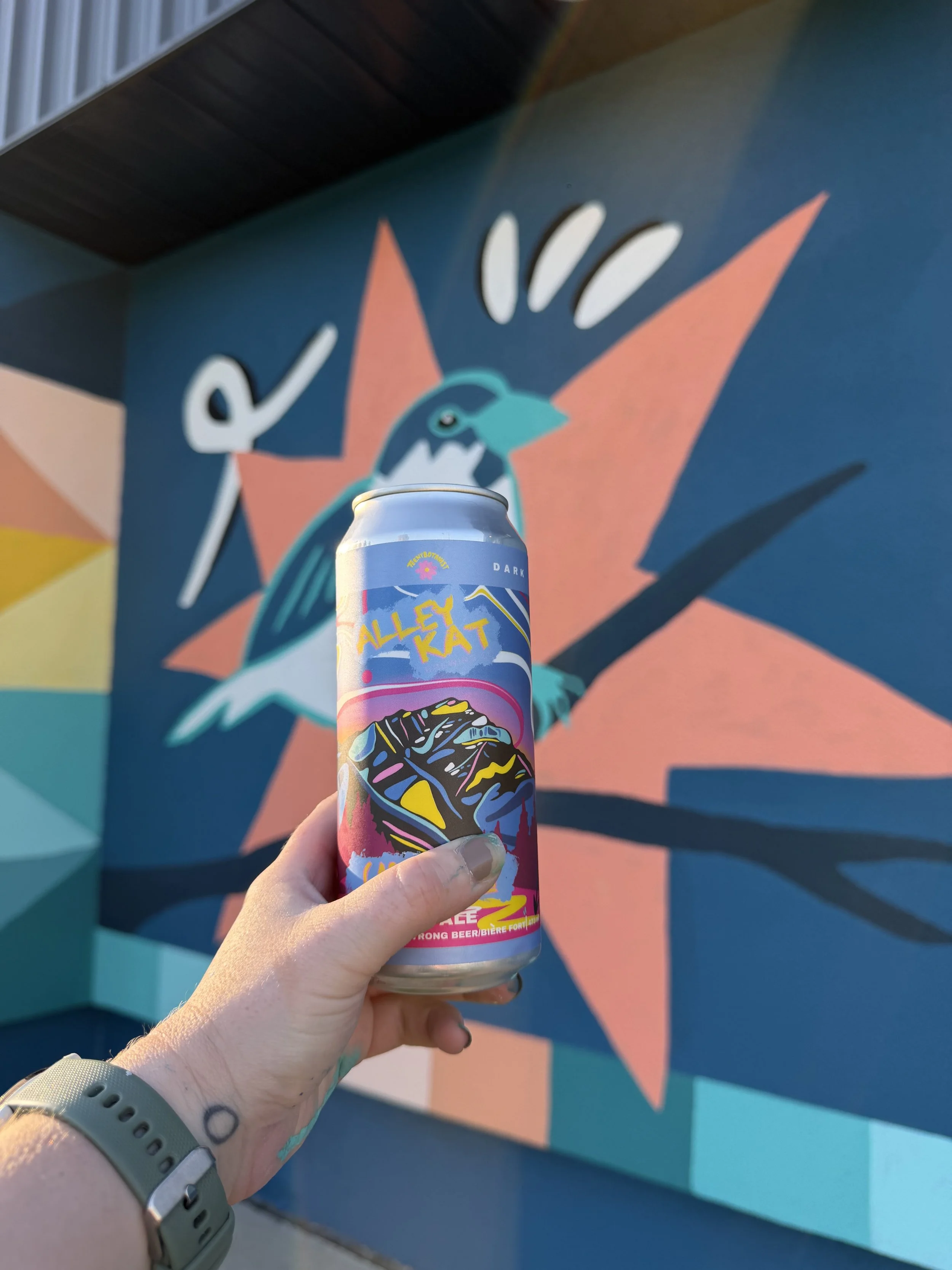 Alley Kat Cascadian Dark Ale can held against a colourful mural
