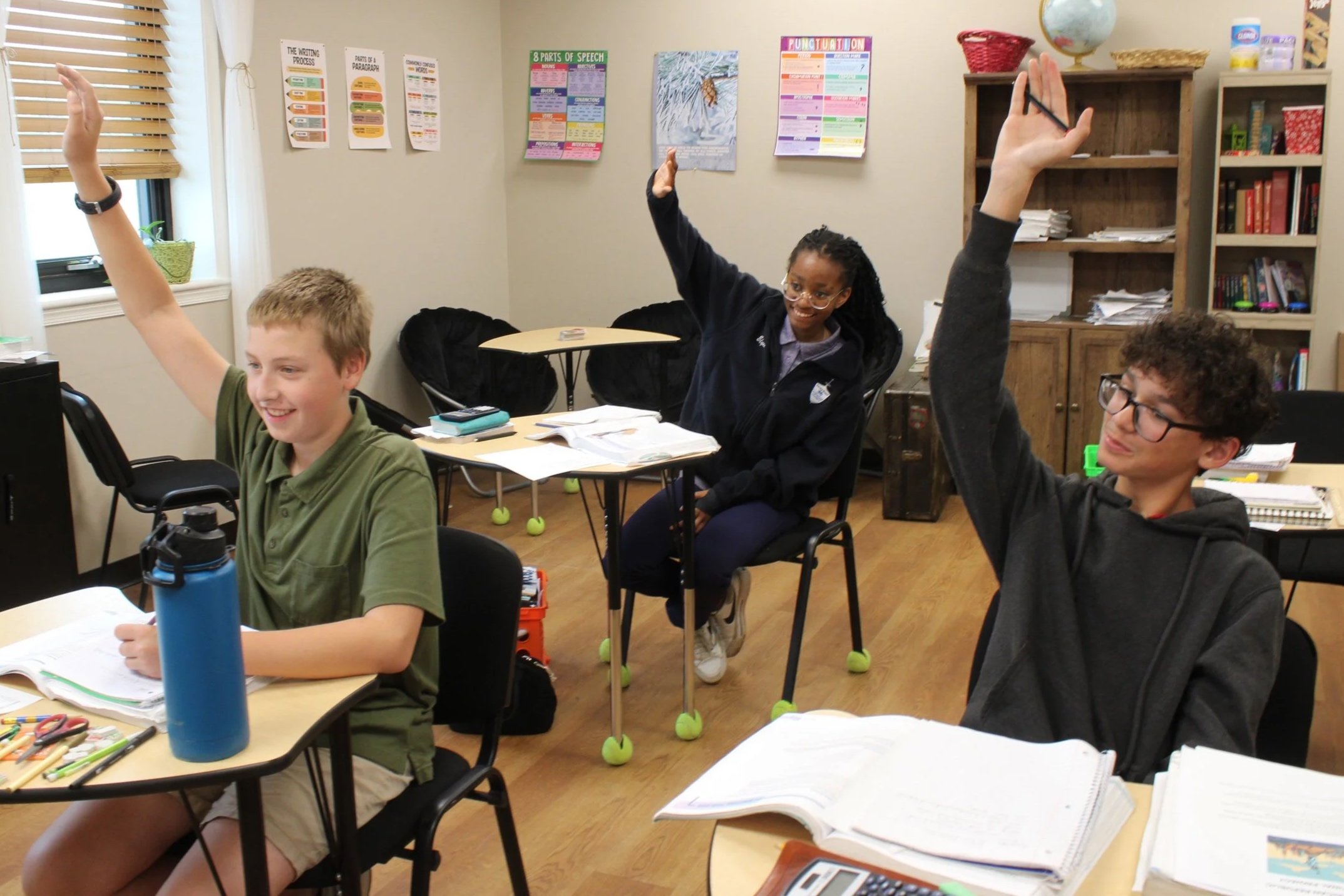Students in a classroom with three raising their hands. The classroom has educational posters on the wall and shelves with books and supplies.