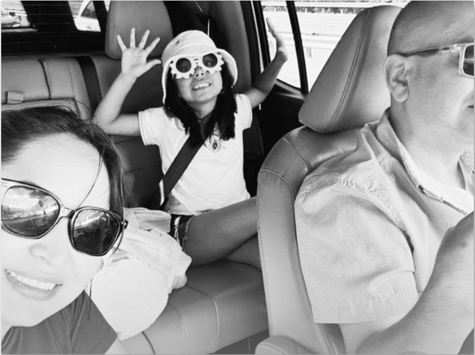 A woman wearing large sunglasses smiling, a young girl in sunglasses and a hat smiling, and a man with glasses, all sitting in a car.