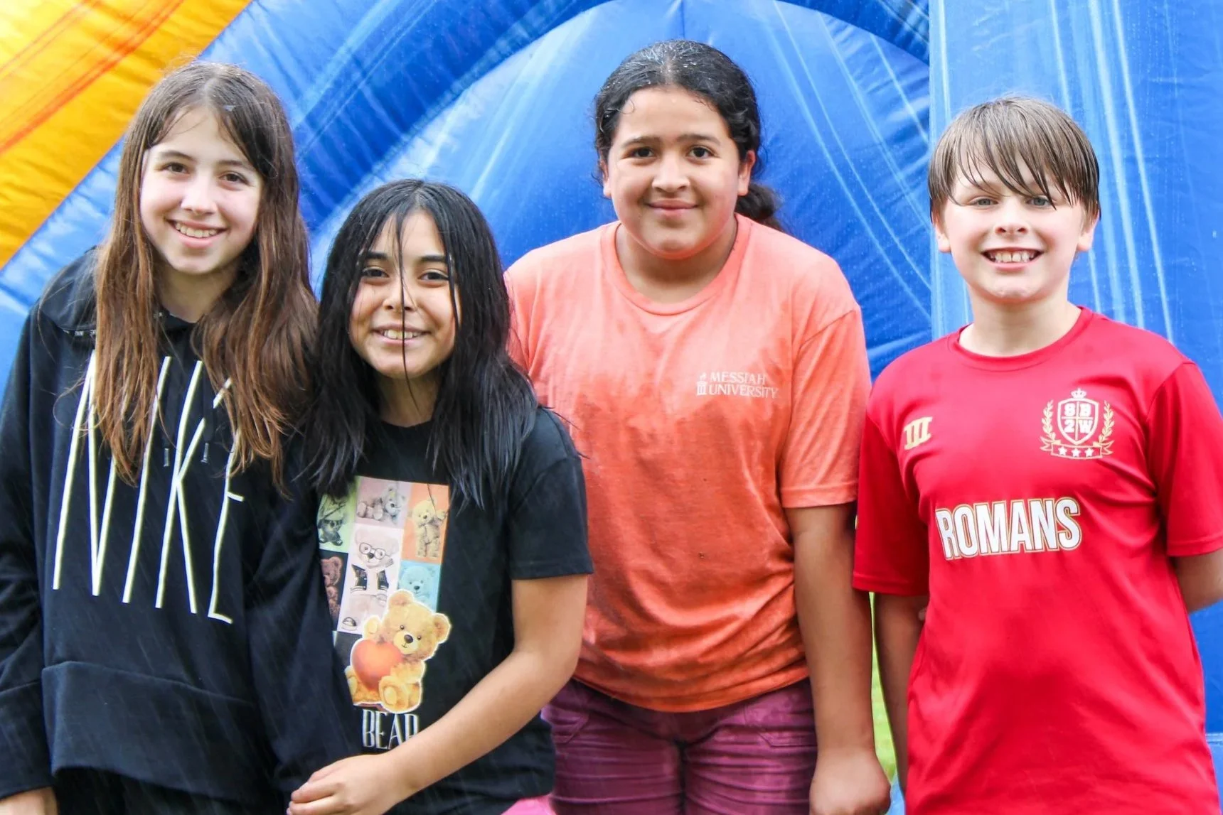 Four children standing outside, smiling at the camera, with a colorful blue, yellow, and orange arch tent in the background.