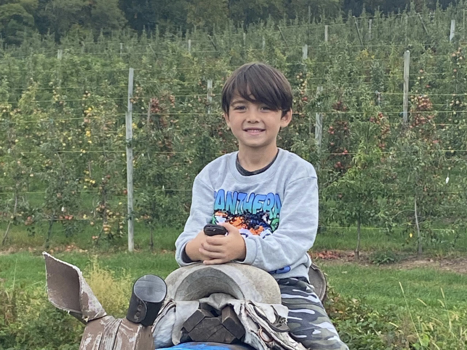A young boy sitting outdoors in front of a lush green orchard, smiling and holding a device, with a camera and a bag in front of him.